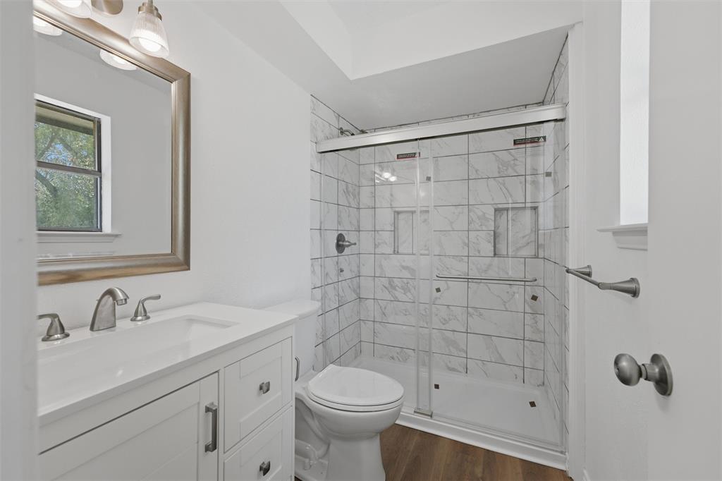 124 Prairie Creek Road Red Oak, TX 75154 - Photo 15 of 17 Spa-inspired ensuite bathroom featuring a walk-in shower with subway tile surround and frameless glass enclosure, white vanity with dual-handle faucet and undermount sink, white quartz countertop, toilet with chrome trim, wood-finish flooring, and framed mirror with integrated lighting