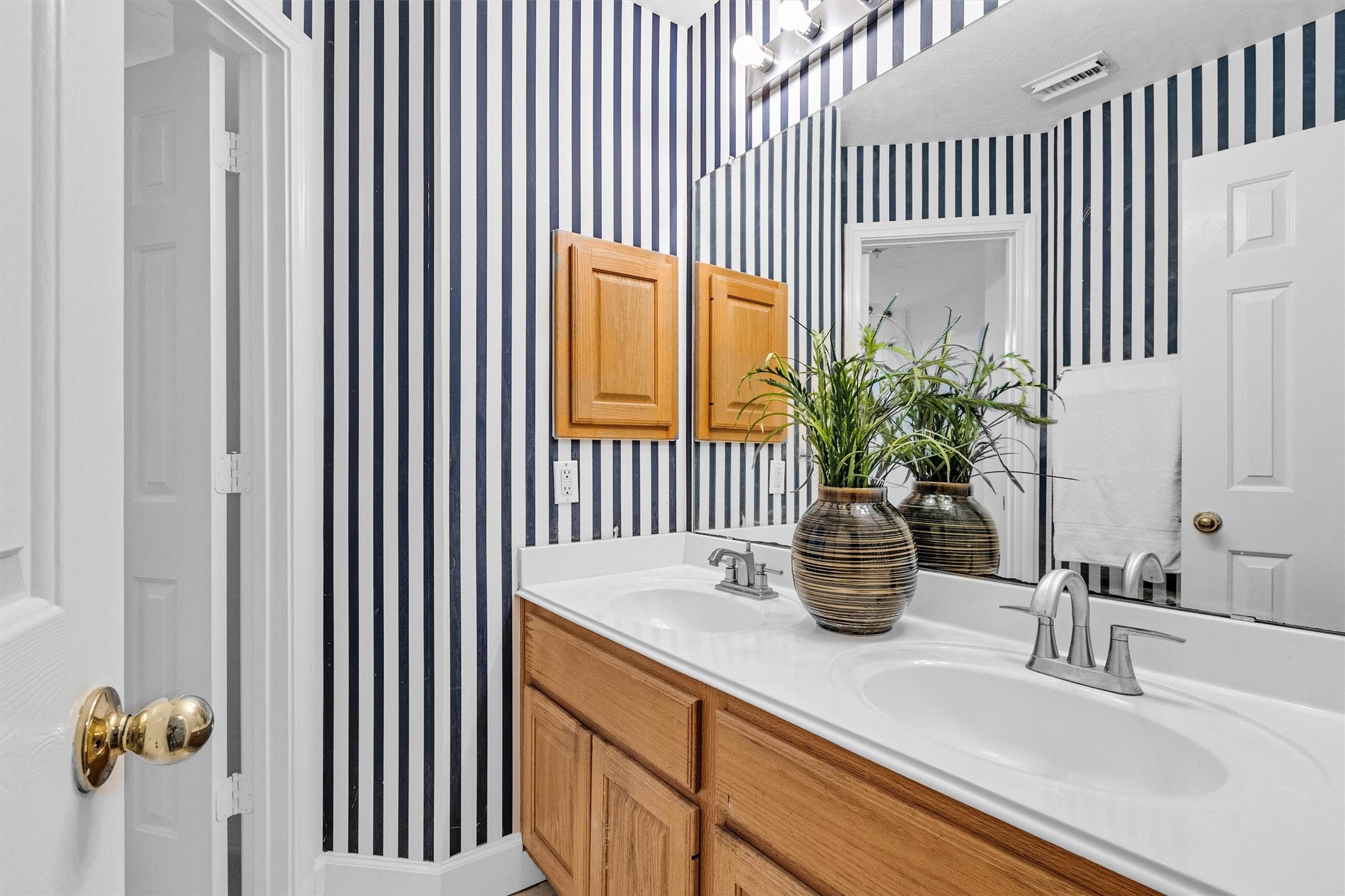 23 Alba Road Montgomery, TX 77356 - Photo 27 of 37 This bathroom features stylish black and white striped wallpaper, a double vanity with a large mirror, wooden cabinetry, and modern fixtures. A decorative plant adds a touch of greenery.