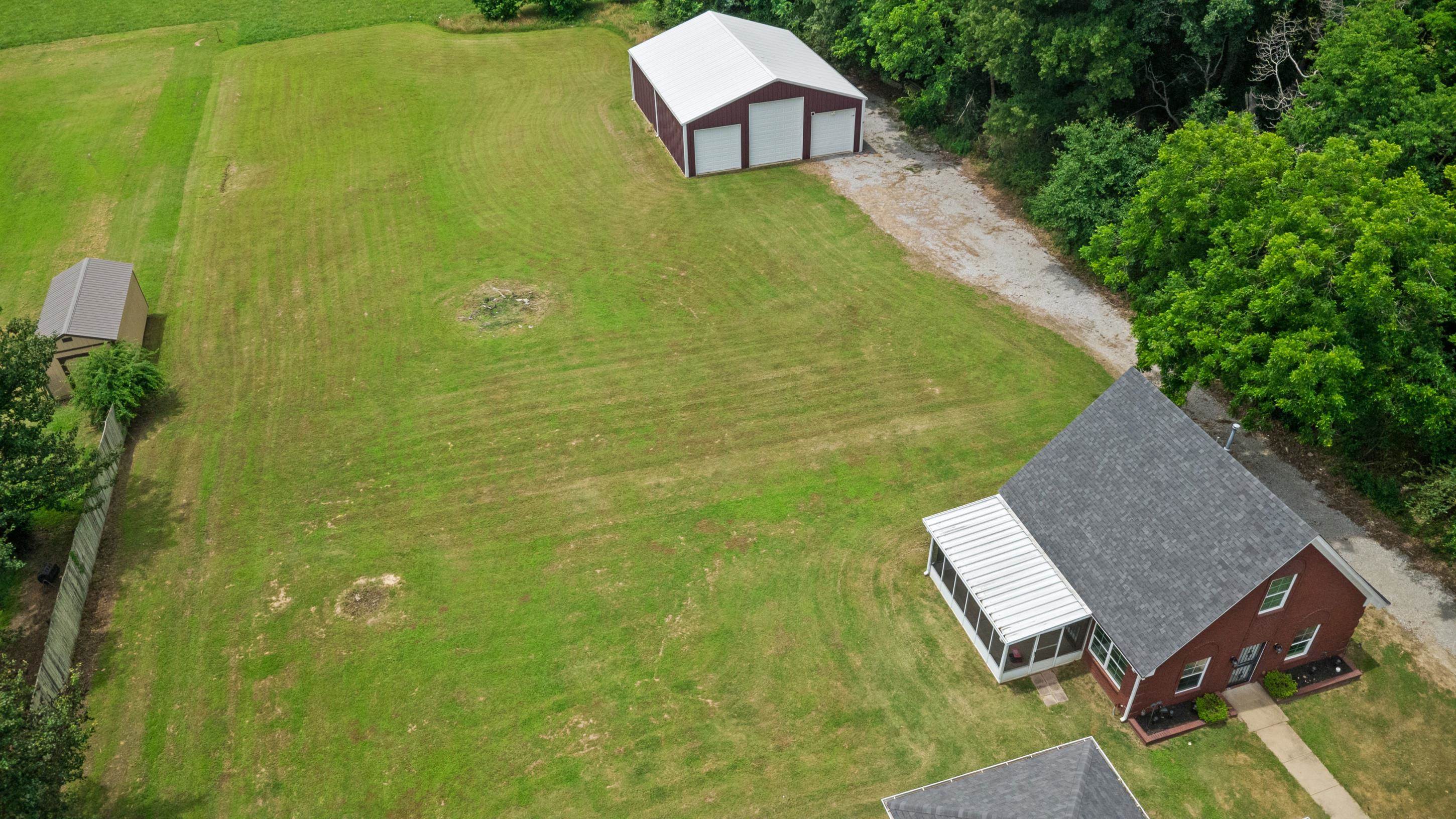 7757 Brunswick Road Millington, TN 38053 - Photo 40 of 40 Expansive green landscape featuring a red brick residence with a gray shingle roof, an attached screened porch, a detached metal barn with three bays, and a secondary shed structure