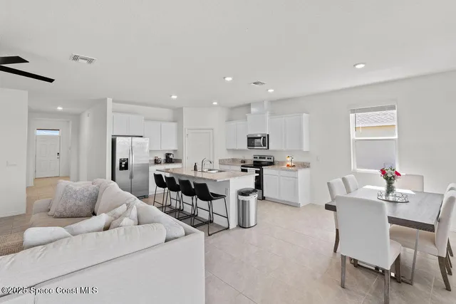 a living room with couches a kitchen island with white cabinets and stainless steel appliances