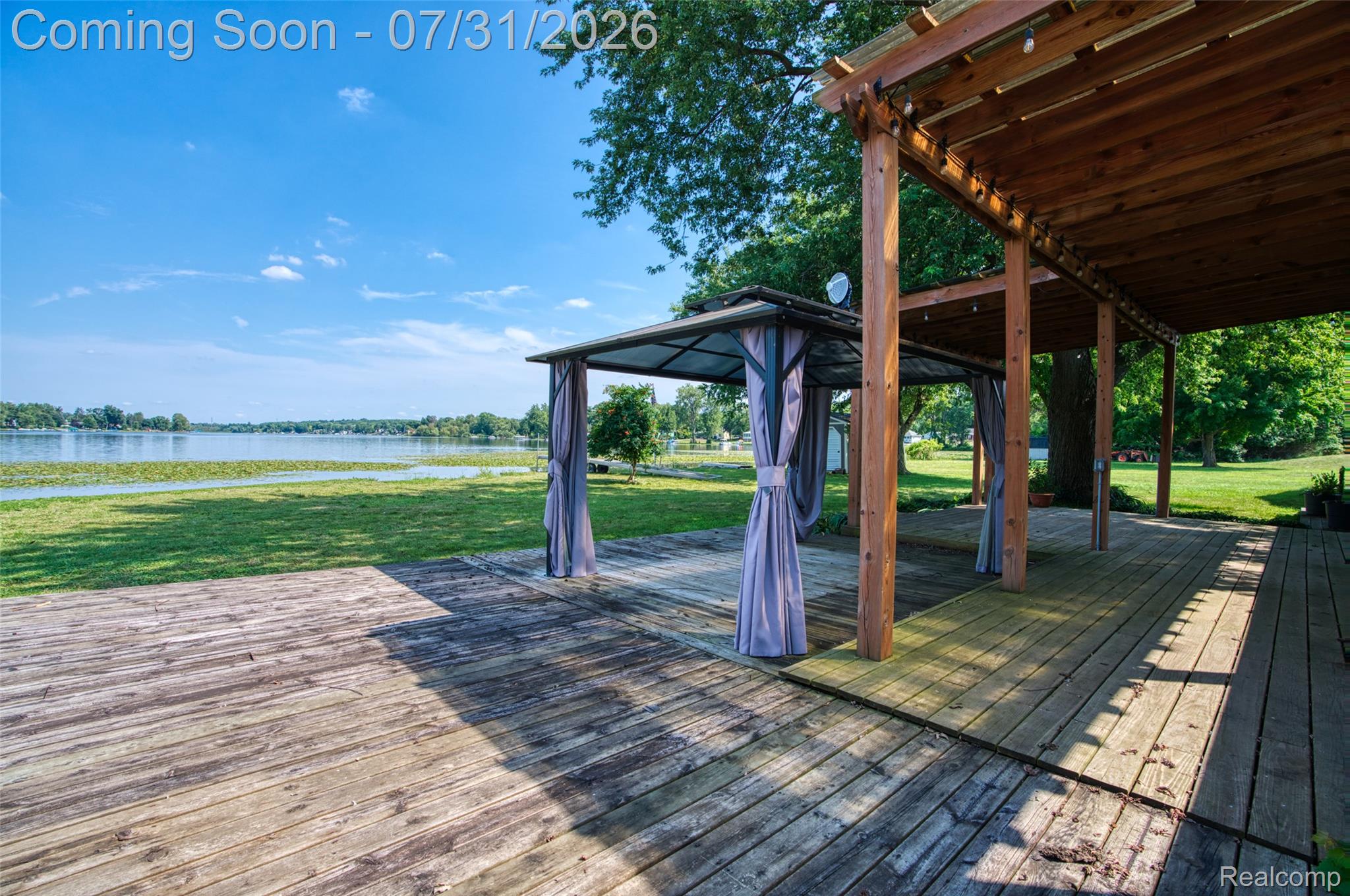 5269 Reno Road Manchester, MI 48158 - Photo 31 of 64 Expansive wood-finish decking featuring a covered pergola and a freestanding gazebo with draped side panels, providing direct access to the waterfront and manicured lawn