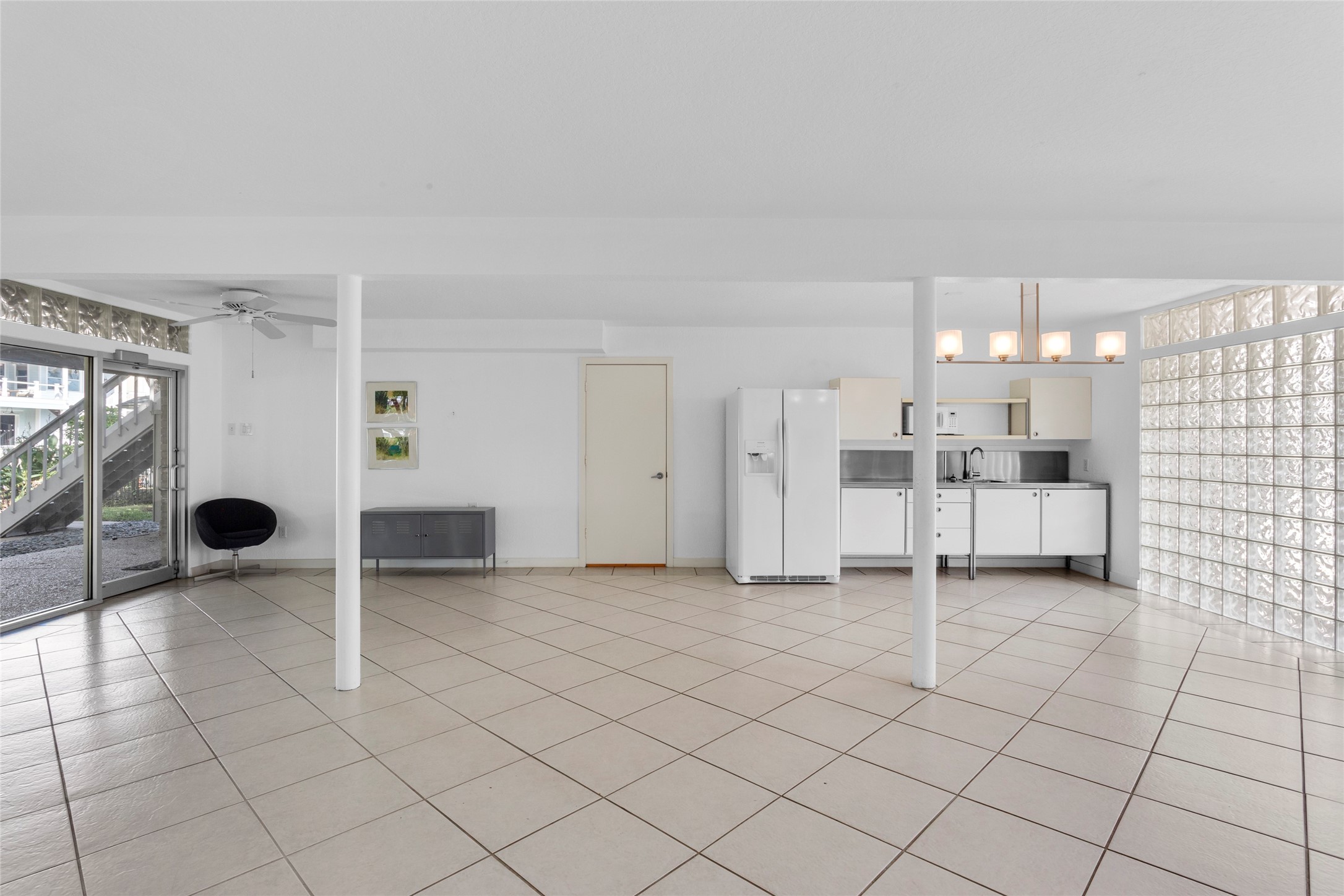67 Harbor Lane Kemah, TX 77565 - Photo 27 of 47 This spacious room features large tile flooring, a minimalist kitchen with white cabinetry, and a bright atmosphere enhanced by glass block walls. It includes a ceiling fan, glass doors leading outside, and modern lighting fixtures, offering a versatile open layout.