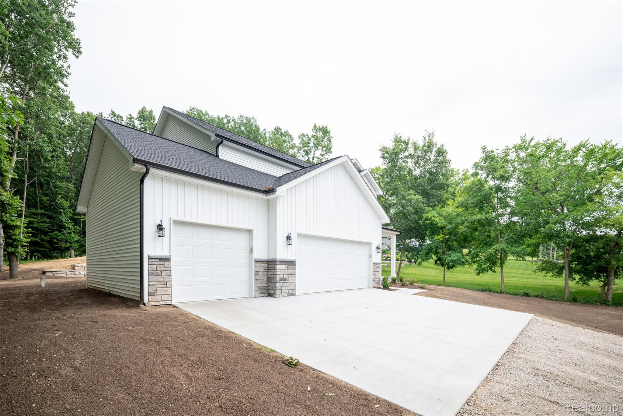 16085 Catalpa Ridge Holly, MI 48442 - Photo 28 of 40 Modern farmhouse-style exterior featuring white vertical siding, a dark shingle roof, two garage bays with white doors, stone veneer accents, and exterior lighting fixtures