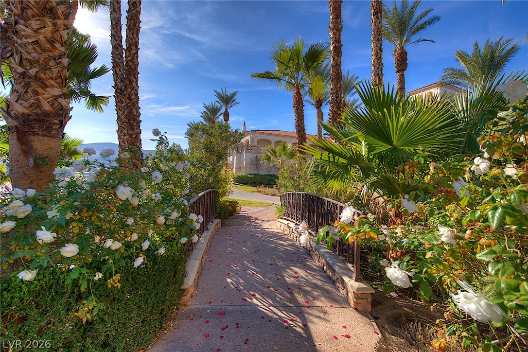 25 Falling Ridge Lane Henderson, NV 89011 - Photo 44 of 48 An inviting stone bridge spans over a lush garden, leading to a charming property under a clear sky.