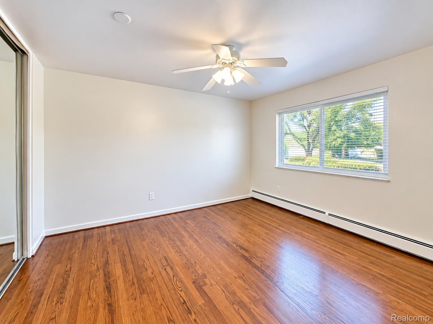 475 South Adams Road, Unit 8 Birmingham, MI 48009 - Photo 8 of 15 Hardwood-style flooring, white ceiling fan with integrated lighting, large window with blinds, baseboard heating, and a mirrored closet door