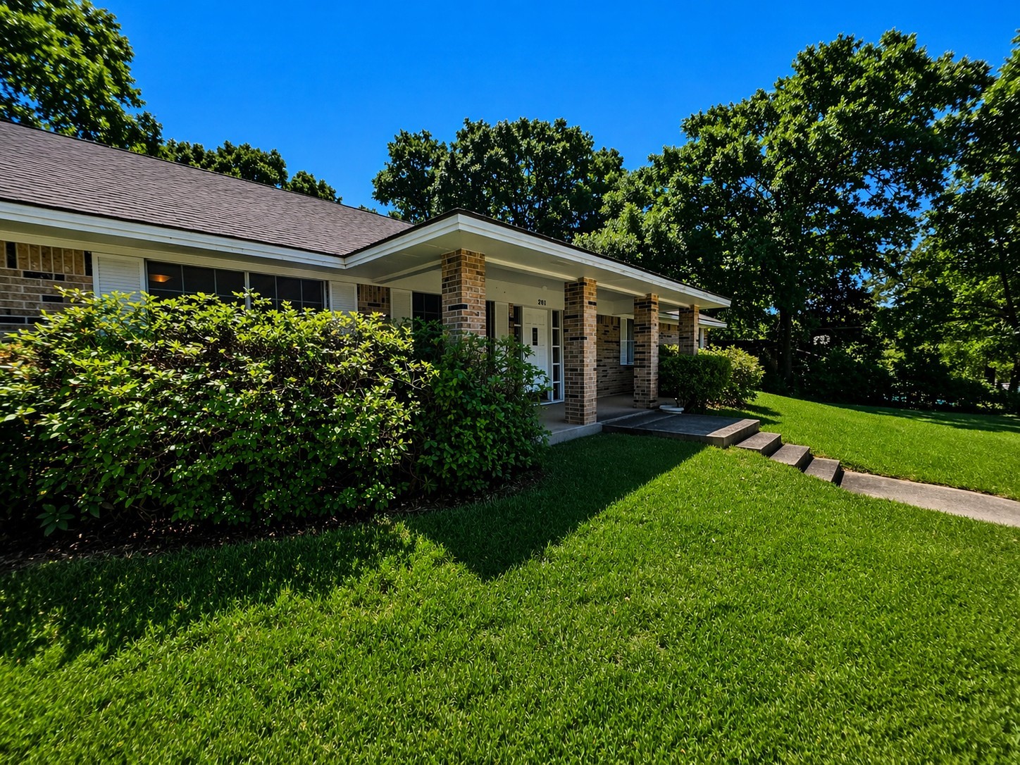 201 Verna Street Jasper, TX 75951 - Photo 22 of 24 This photo showcases a charming, single-story brick house with a welcoming front porch. It is surrounded by well-maintained greenery and mature trees, enhancing its curb appeal. The lawn is lush and the setting is serene, perfect for a cozy home environment.