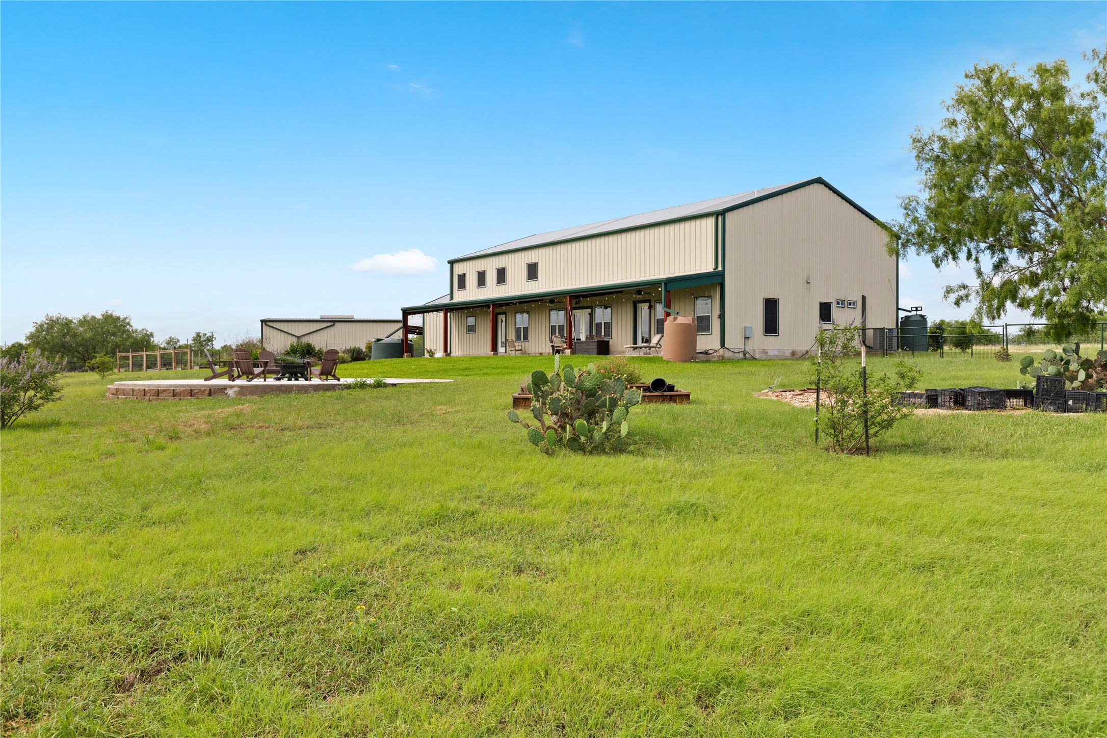 1293 Pioneer Road Seguin, TX 78155 - Photo 5 of 40 An inviting covered patio and fire pit area extend from this broad, light-colored structure, set against a vibrant green landscape.