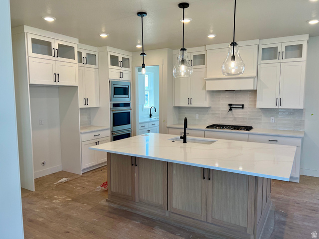 11507 Markfield Way, Unit 1124 South Jordan, UT 84009 - Photo 2 of 6 Contemporary kitchen featuring a large central island with a sink, white cabinetry with black hardware, stainless steel double wall ovens, a gas range with a pot filler, and pendant lighting