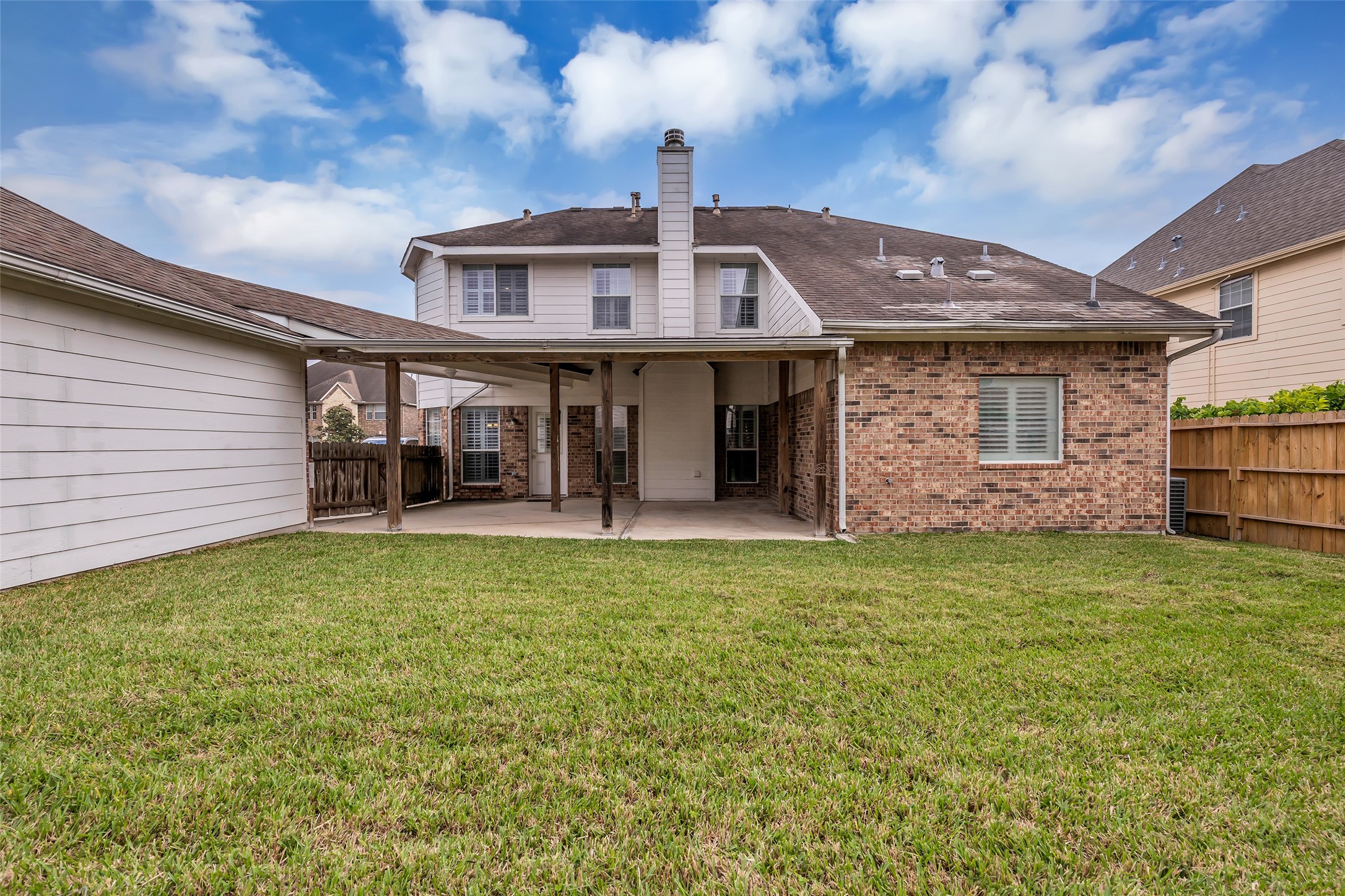 6807 Chapelfield Lane Houston, TX 77049 - Photo 41 of 42 This photo showcases the backyard of a two-story home with a mix of brick and siding exterior. It features a covered patio, providing a shaded outdoor area, and a well-maintained grassy yard, enclosed by a wooden fence for privacy. Ideal for outdoor activities and relaxation.