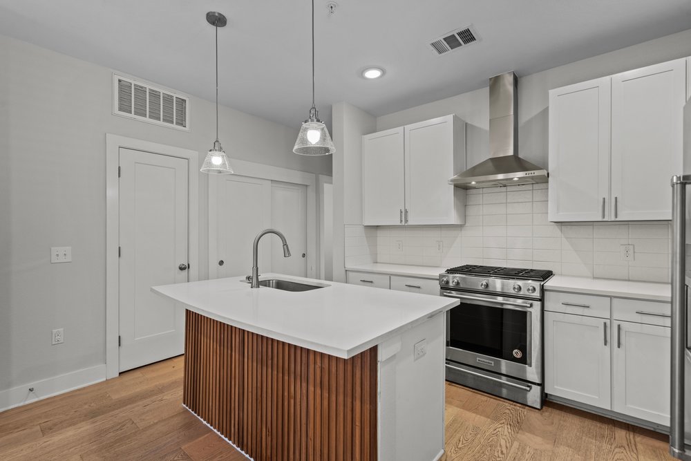 2827 Manor Road, Unit 2201 Austin, TX 78722 - Photo 10 of 40 Kitchen featuring a central island with wood paneling and a stainless steel sink, under-cabinet lighting, wood-finish flooring, white flat-panel cabinetry, and a subway tile backsplash