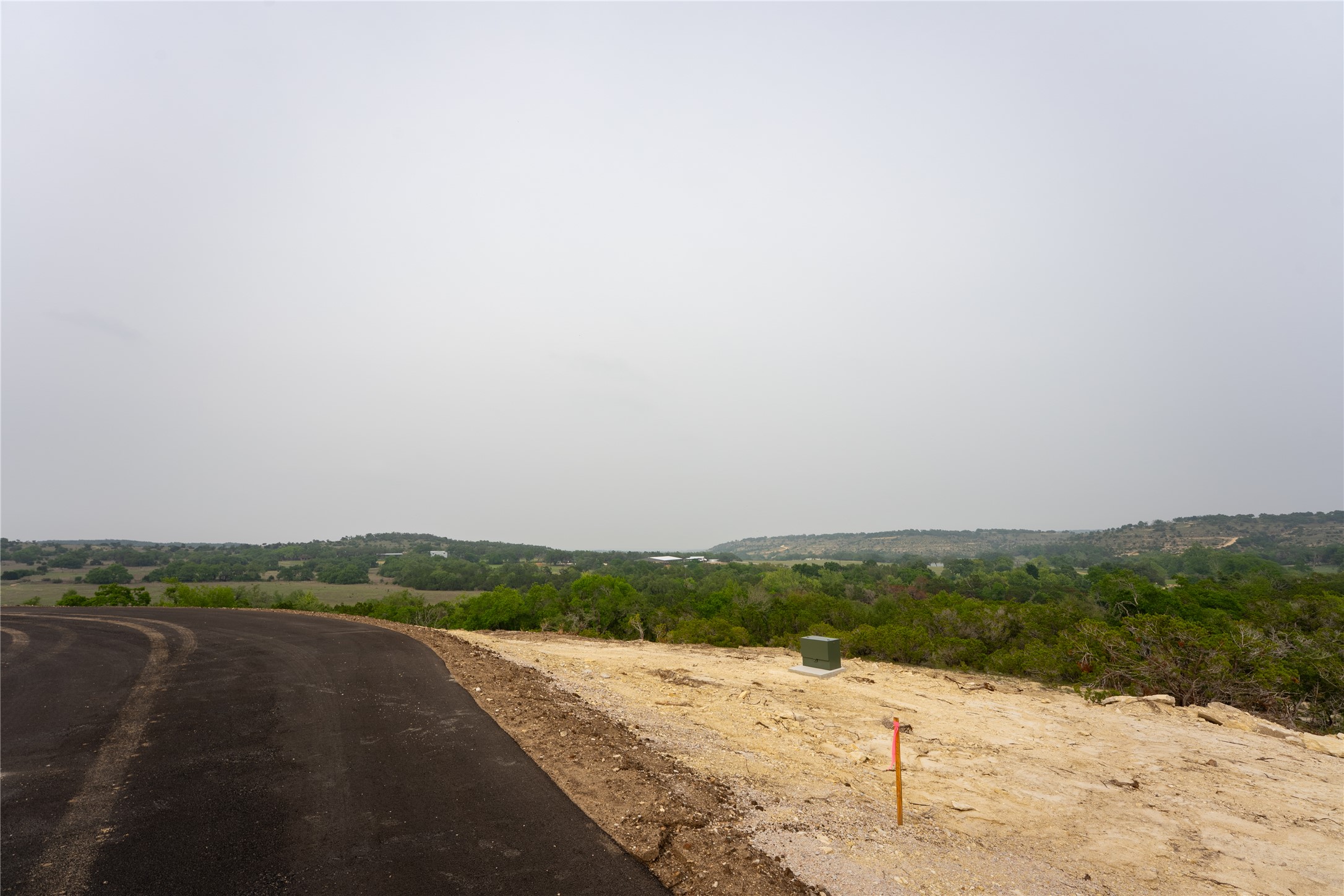 Lot 25 Ranch Road 165 Dripping Springs, TX 78620 - Photo 7 of 8 Paved road bordering a cleared building site, featuring a utility box and distant tree-lined landscape