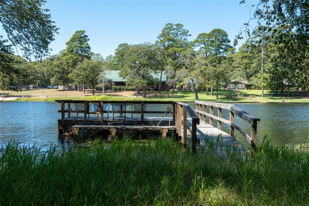 167 Blue Water Point Holly Lake Ranch, TX 75765 - Photo 25 of 27 Wood-plank dock with a built-in seating area over a body of water, surrounded by mature trees and a distant building with a green roof