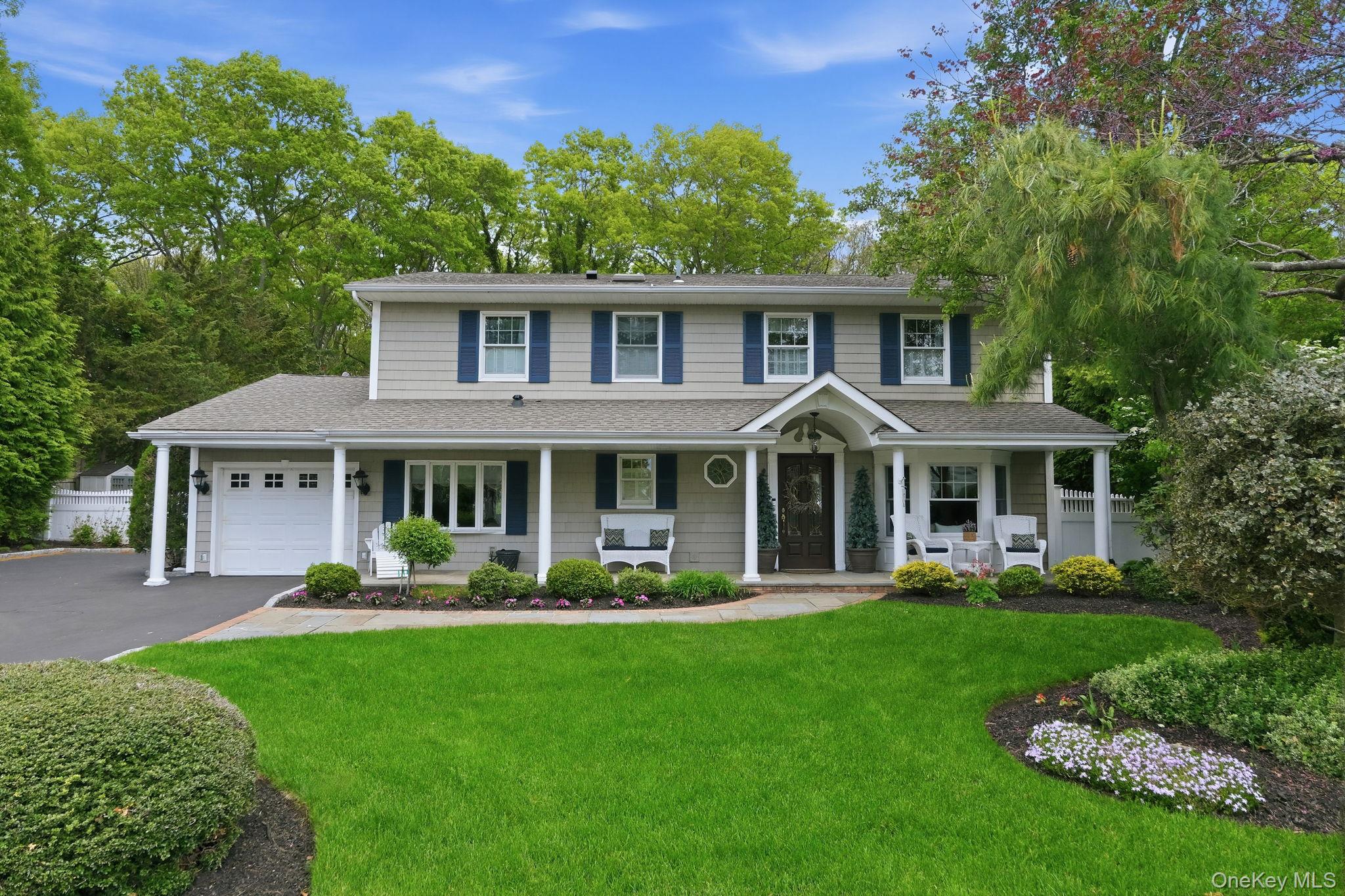 17 Milburn Road Centereach, NY 11720 - Photo 1 of 1 Two-story residence featuring a covered front porch with white columns, a garage, a paved driveway, and established landscaping with mature trees and shrubs