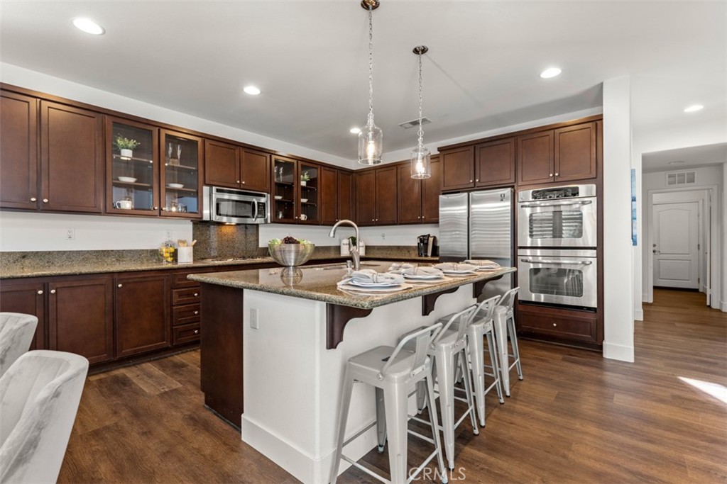 14977 Brooktree Street Eastvale, CA 92880 - Photo 6 of 34 a kitchen with stainless steel appliances granite countertop a kitchen island a stove a refrigerator a sink a dining table and chairs with wooden floor
