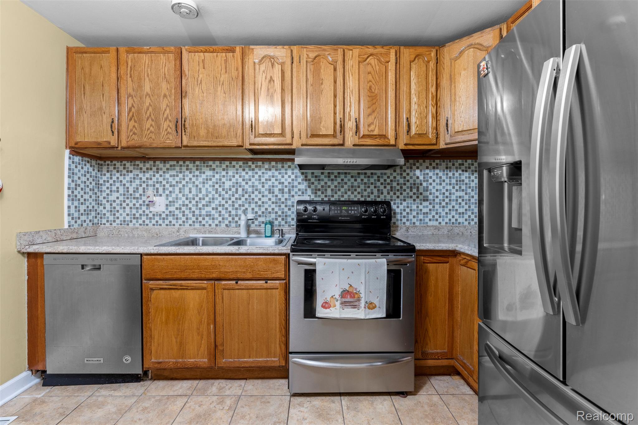 22530 Sheridan Road Novi, MI 48375 - Photo 12 of 50 Kitchen featuring appliances with stainless steel finishes, decorative backsplash, brown cabinets, light tile patterned flooring, and under cabinet range hood