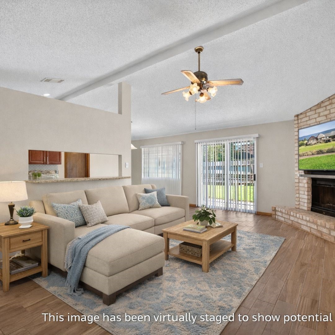 11803 Inga Lane Houston, TX 77064 - Photo 11 of 39 This photo showcases a virtually staged living room with a cozy beige sectional sofa, wooden coffee table, and side table. The room features a ceiling fan, large windows with vertical blinds for natural light, and a brick fireplace. Hardwood flooring and neutral walls create a warm, inviting space.