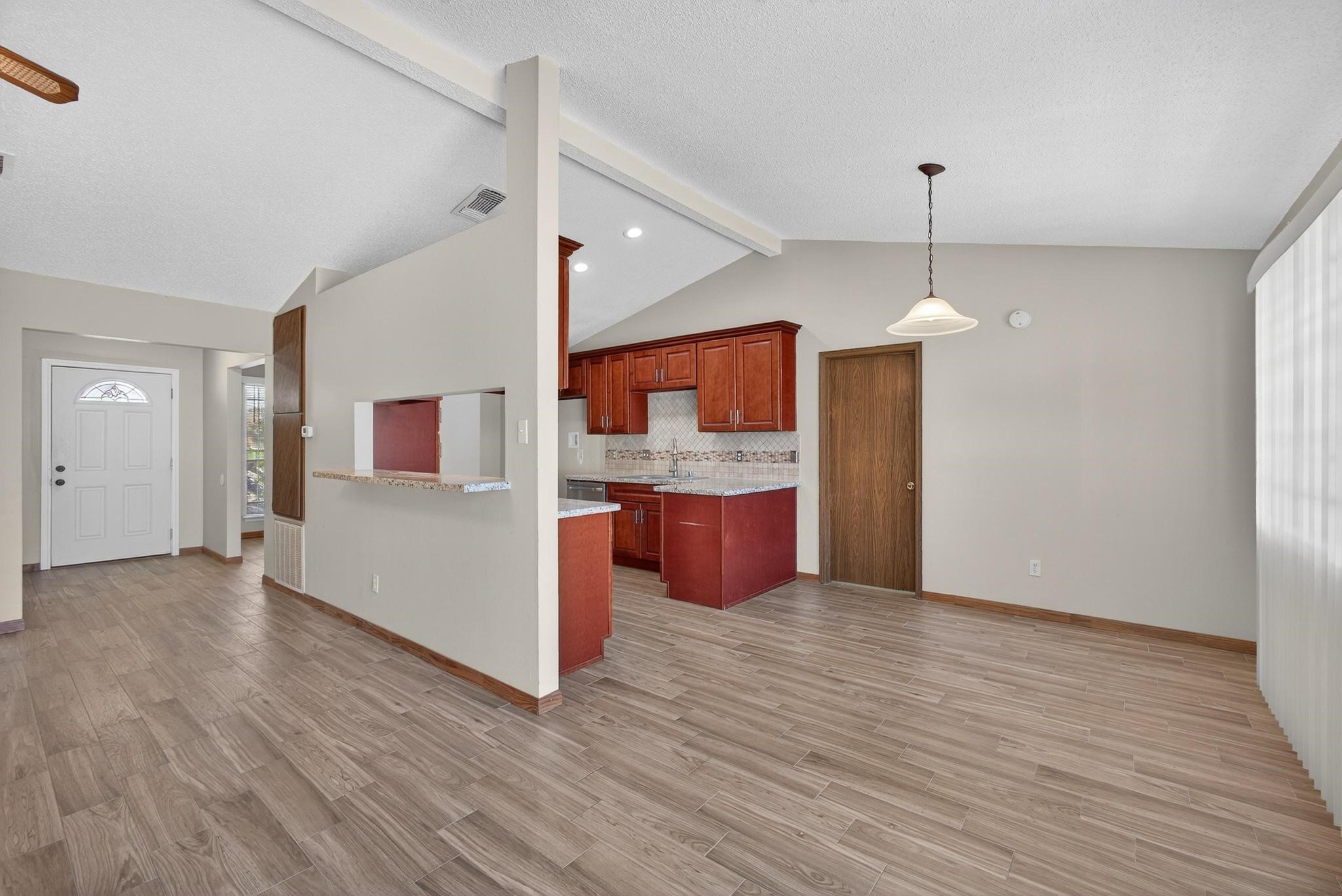 11803 Inga Lane Houston, TX 77064 - Photo 14 of 39 This photo showcases an open-concept living space with wood-look flooring and neutral walls. It features a modern kitchen with cherry wood cabinets, a tiled backsplash, and a breakfast bar. The dining area has a pendant light, and natural light fills the room through large windows with vertical blinds.