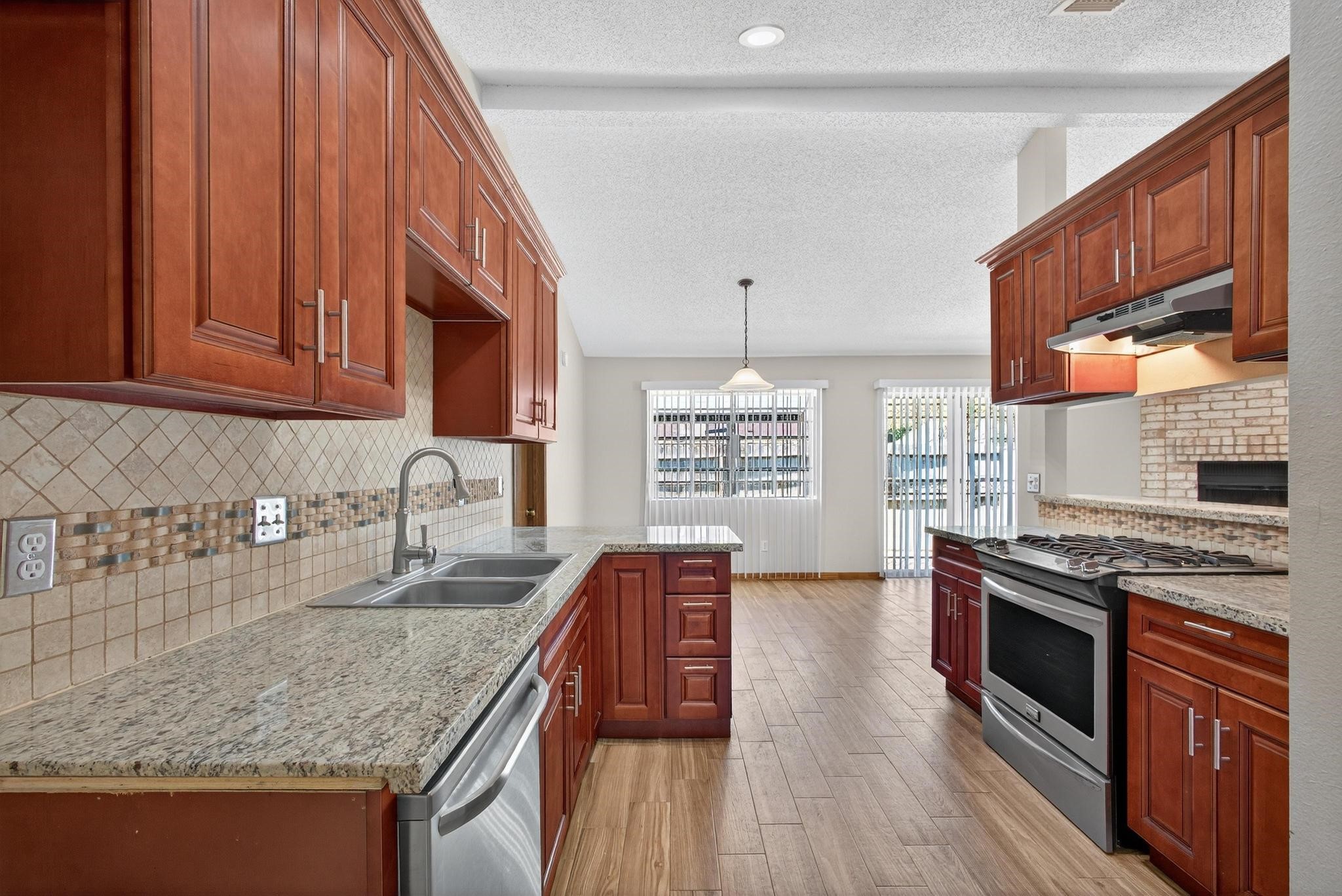 11803 Inga Lane Houston, TX 77064 - Photo 17 of 39 This kitchen features elegant wooden cabinetry, granite countertops, and stainless steel appliances. It has a tiled backsplash and opens to a bright dining area with large windows and wood-style flooring. A cozy fireplace adds charm to the space.