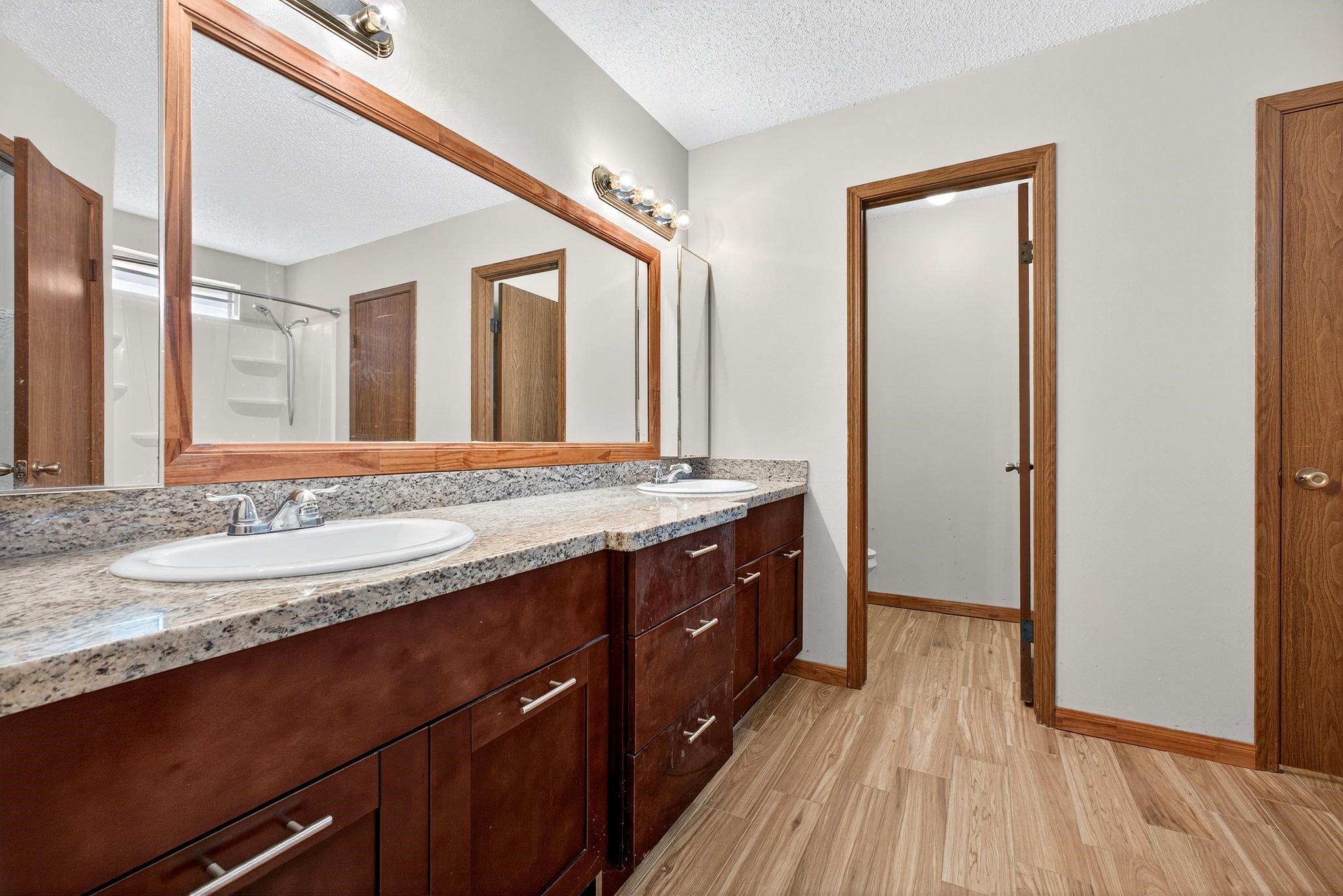 11803 Inga Lane Houston, TX 77064 - Photo 24 of 39 This bathroom features a spacious double vanity with granite countertops and ample storage. It has a large mirror, modern lighting, and a clean, neutral design with wood accents and flooring. A shower area is visible in the background.