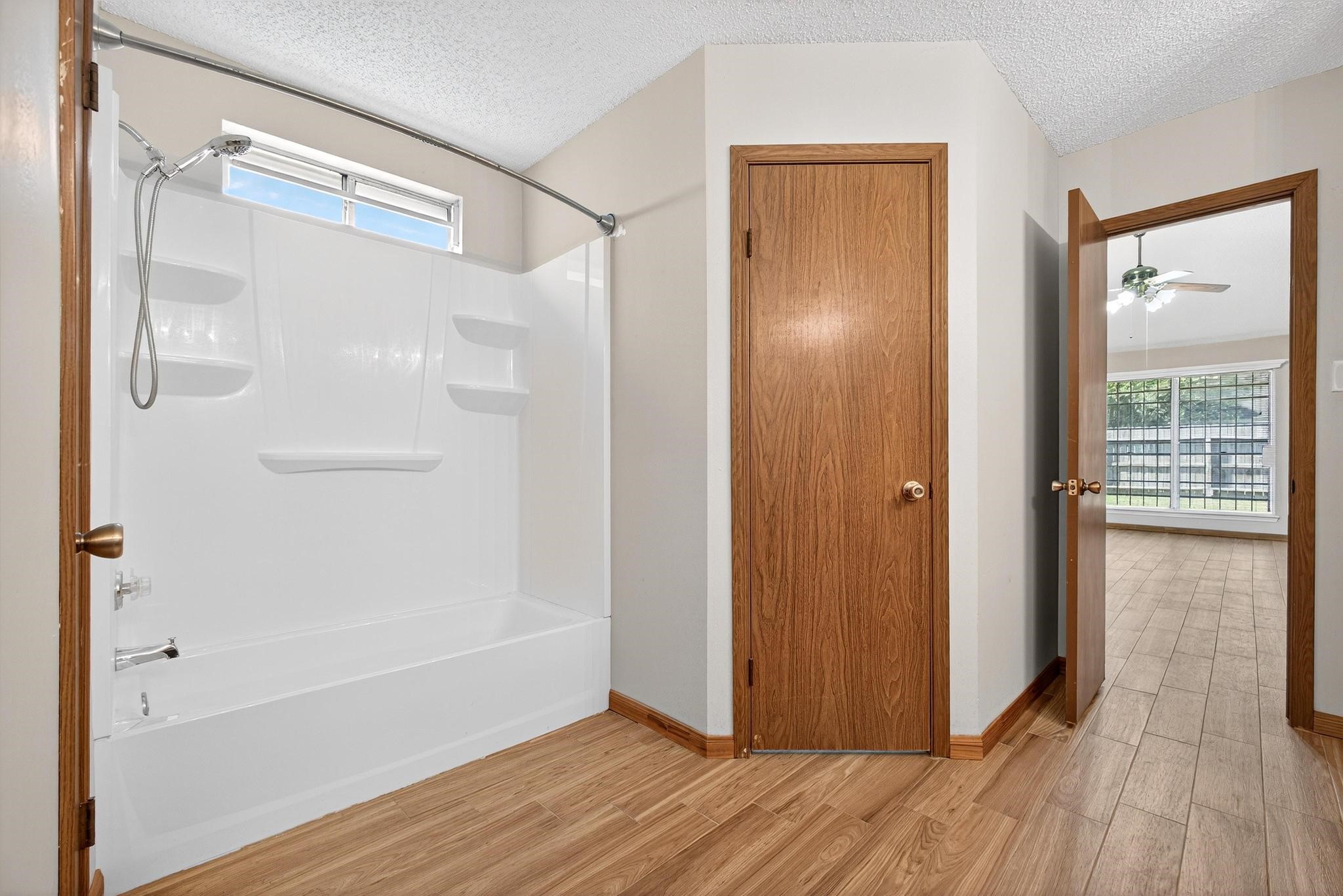 11803 Inga Lane Houston, TX 77064 - Photo 26 of 39 This photo features a bathroom with a white bathtub-shower combo, wood flooring, and two wooden doors. One door leads to a bright room with large windows, providing ample natural light.