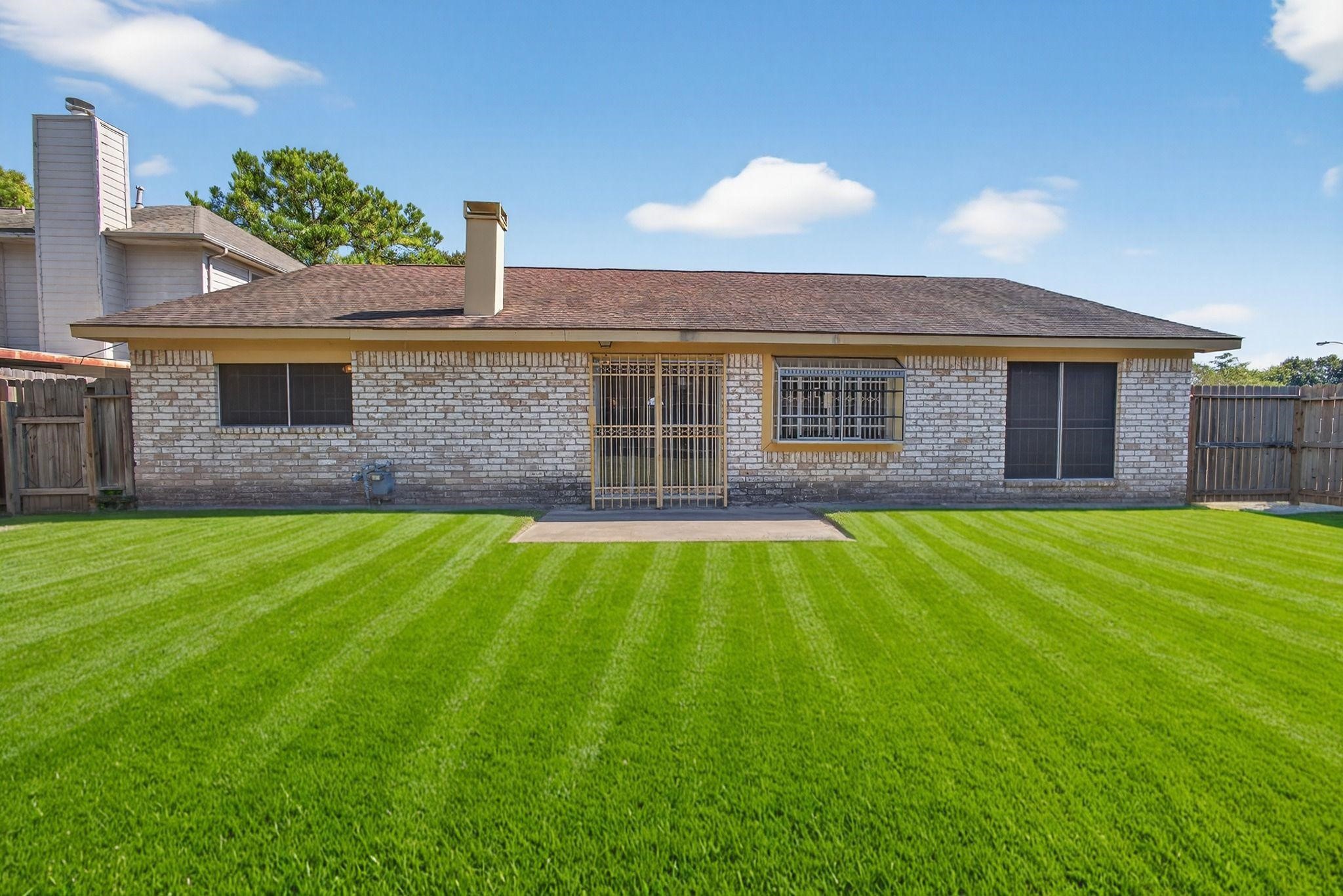 11803 Inga Lane Houston, TX 77064 - Photo 4 of 39 This photo shows the rear view of a single-story, brick house with a well-maintained lawn. The backyard is fenced, featuring a small patio area. The roof is sloped, and there are several windows with security bars.