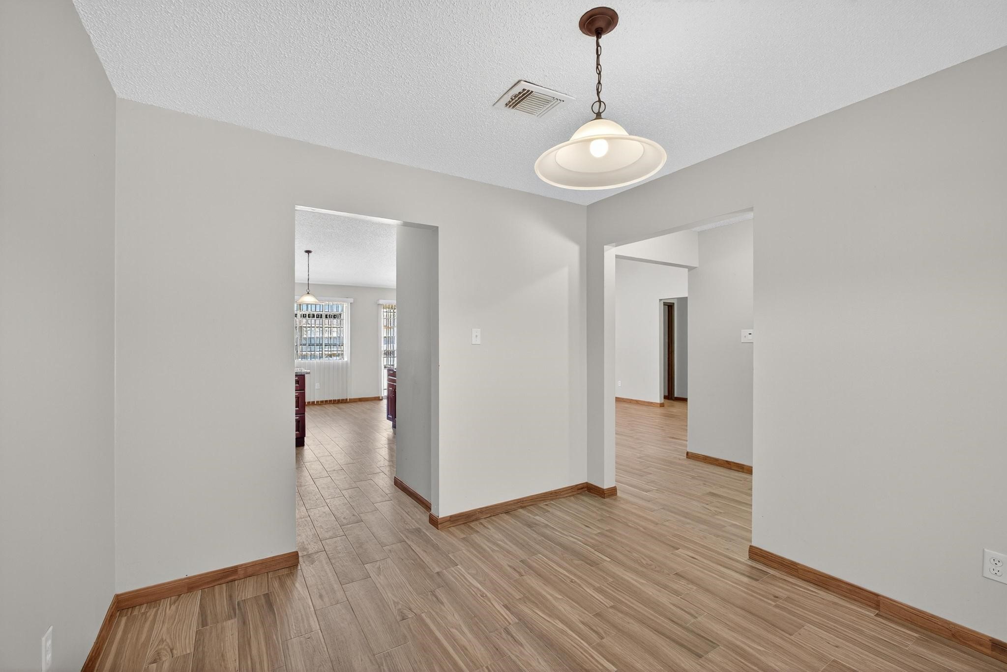 11803 Inga Lane Houston, TX 77064 - Photo 10 of 39 This photo showcases a bright, open hallway with light-colored walls and wood flooring. It features multiple doorways leading to adjacent rooms and a hanging light fixture. Natural light filters in from a window in the connecting room.