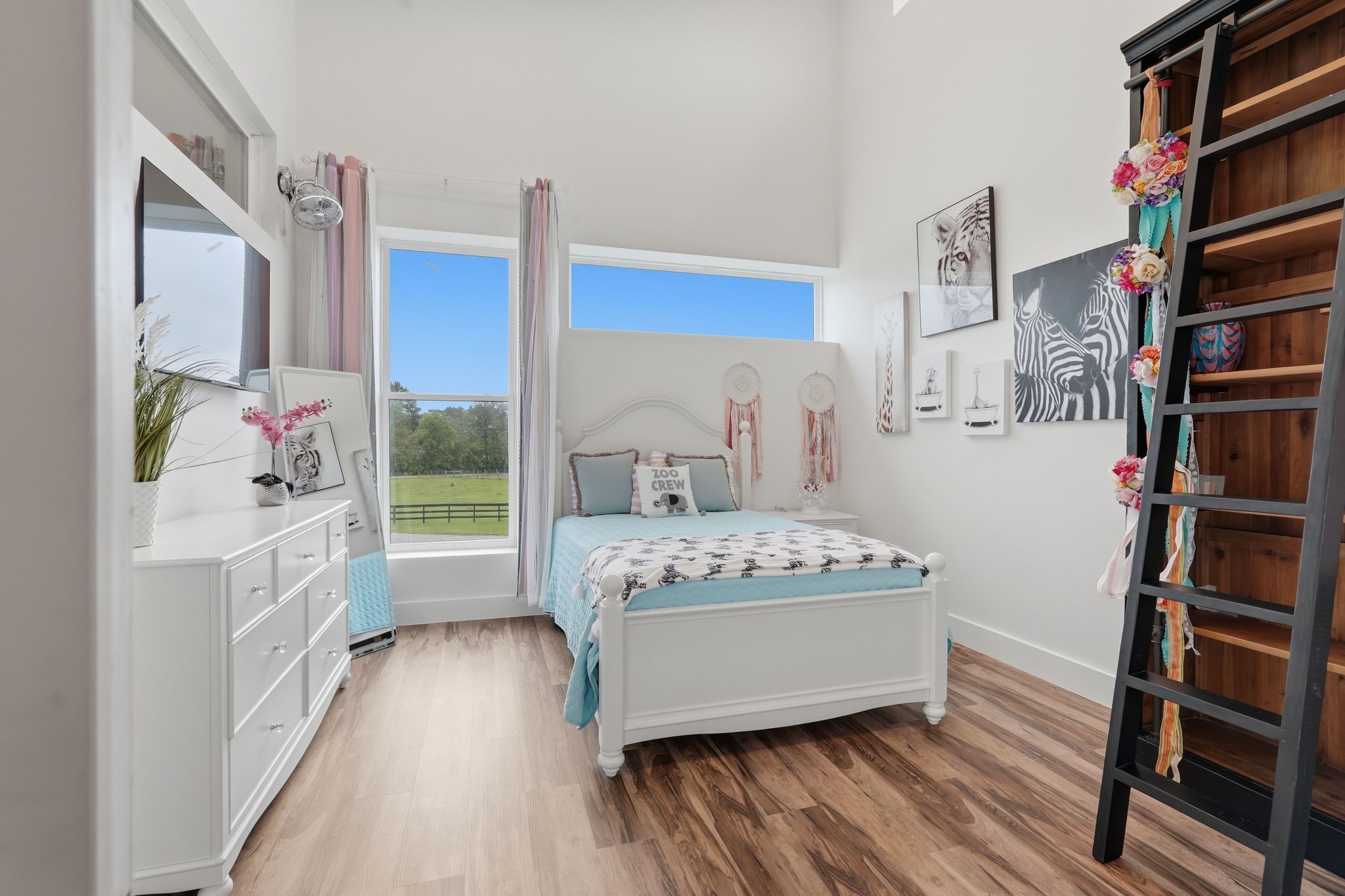 2743 Bobville Road Montgomery, TX 77316 - Photo 34 of 50 Bright and airy bedroom with large windows offering scenic views. Features a cozy bed, stylish decor with zebra artwork, and a ladder leading to a loft area. Warm wood flooring complements the white furniture, creating an inviting atmosphere.