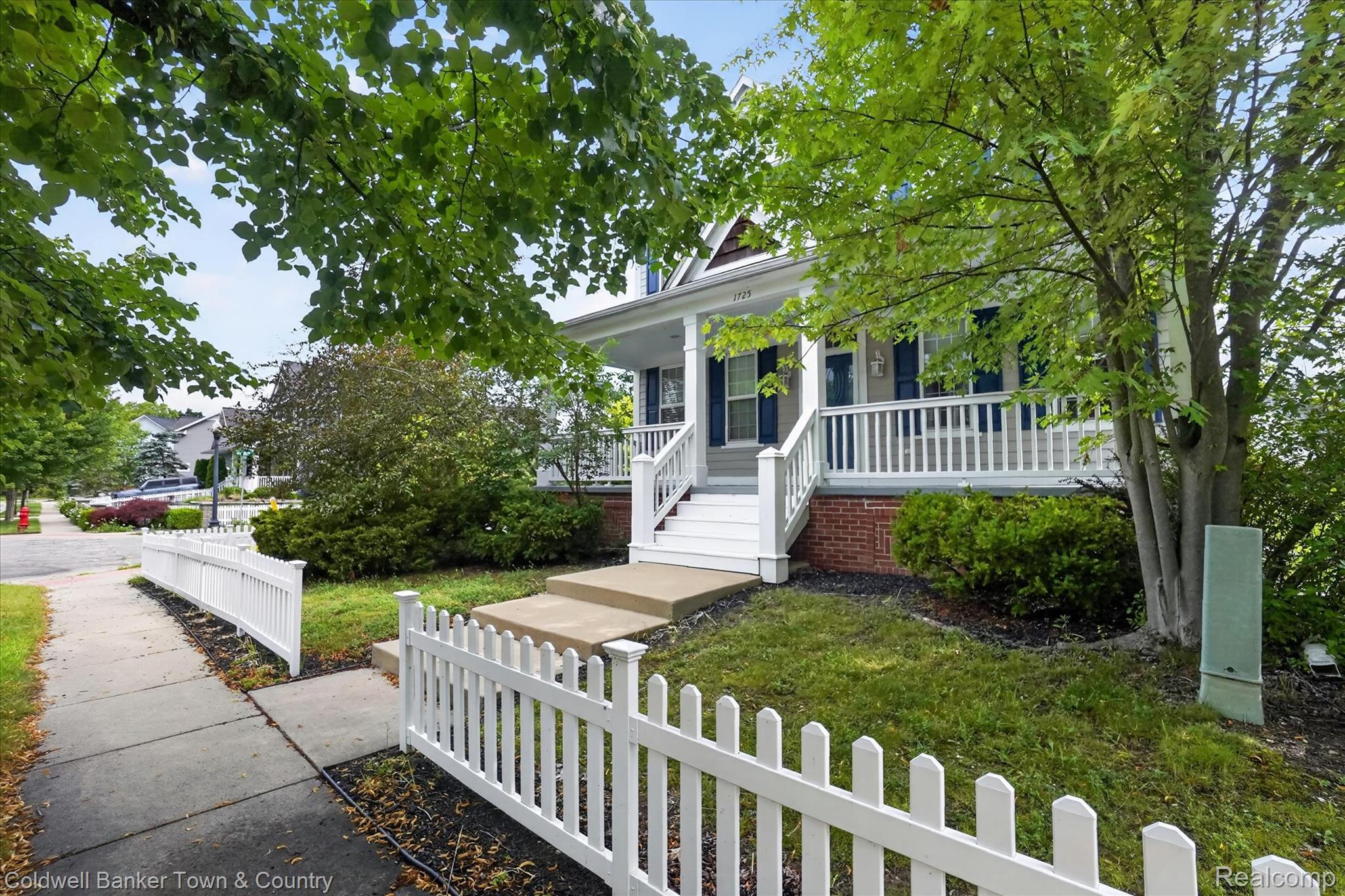 1725 Town Commons Drive Howell, MI 48855 - Photo 5 of 53 Charming exterior featuring a white picket fence, covered front porch with white railings and columns, brick foundation, and mature trees providing natural shade