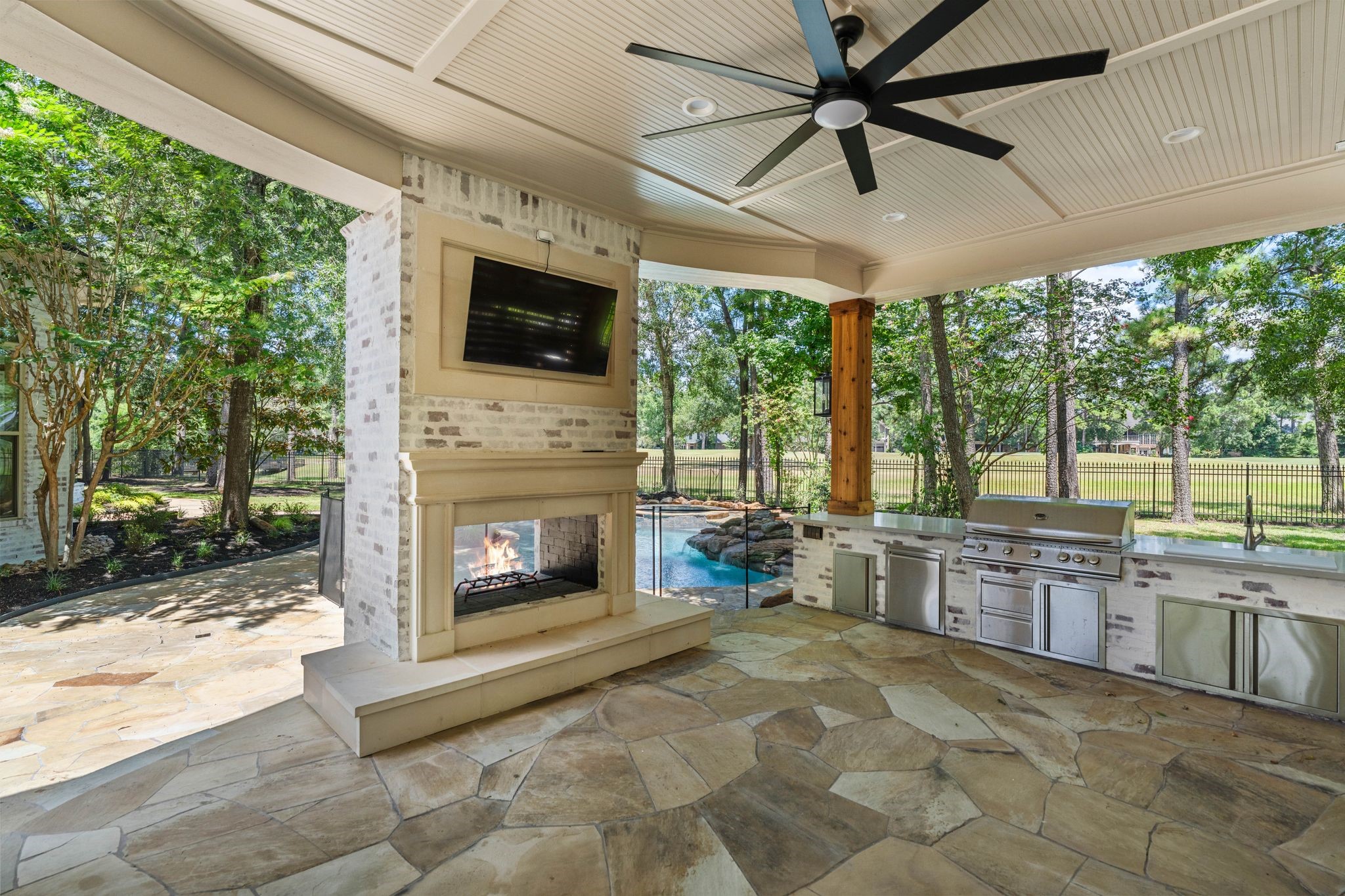 31 Hollymead Drive Spring, TX 77381 - Photo 45 of 50 This photo showcases a luxurious covered outdoor patio featuring a ceiling fan, stone fireplace with a mounted TV, built-in grill and kitchen area, and views of a lush garden and pool, perfect for entertaining.