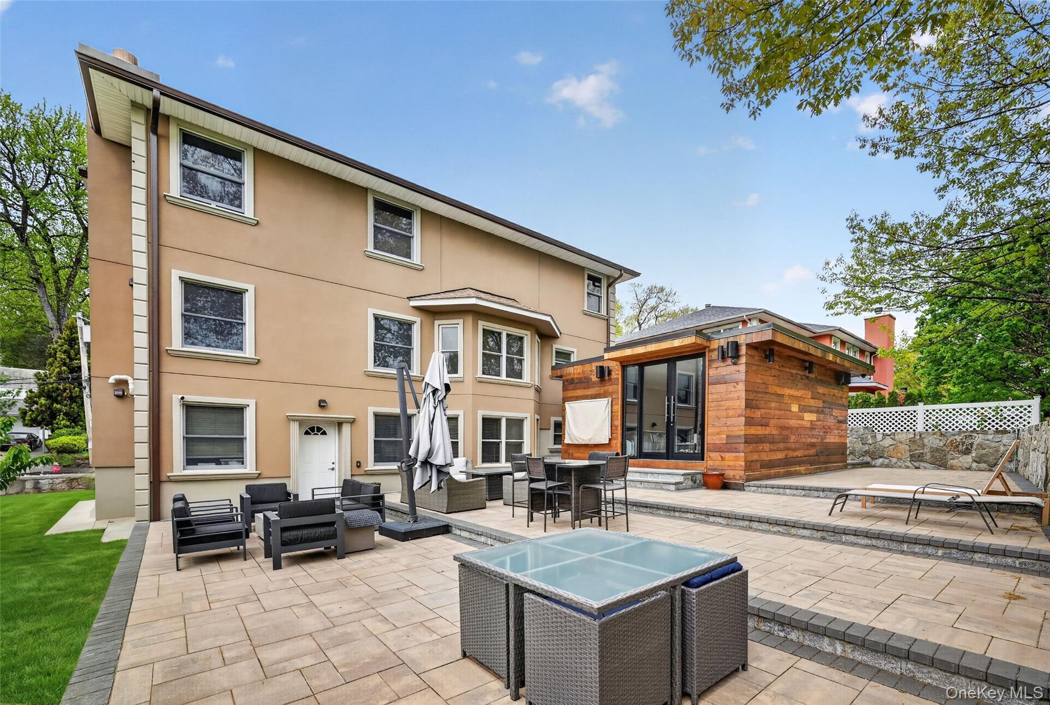 65 Wildway Bronxville, NY 10708 - Photo 20 of 42 Expansive paver patio featuring multiple seating areas, a separate wood-paneled structure with large glass doors, and a stone retaining wall