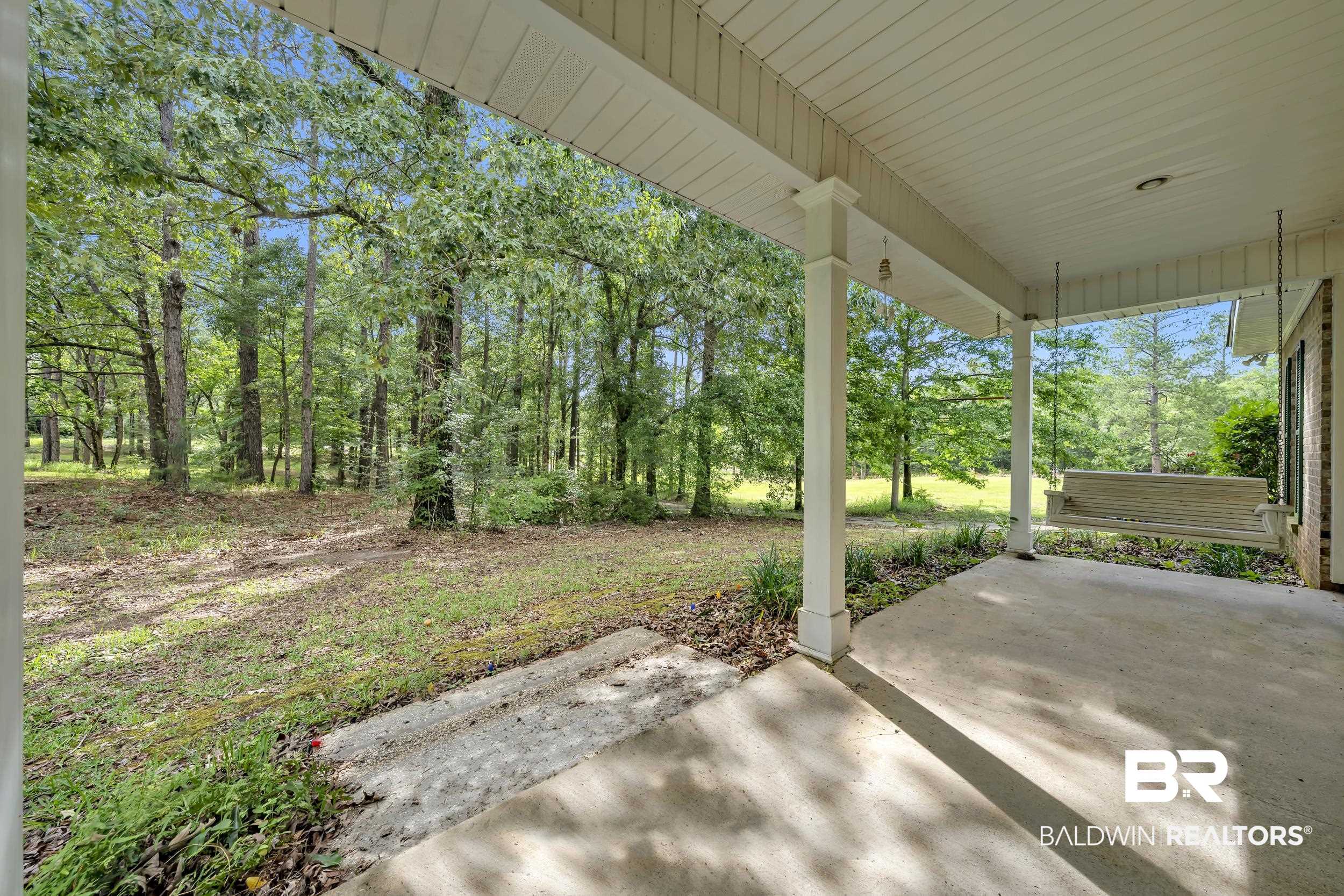 14960 Howells Ferry Road Wilmer, AL 36587 - Photo 3 of 22 Covered porch with white columns, concrete flooring, and a porch swing, overlooking a treed landscape