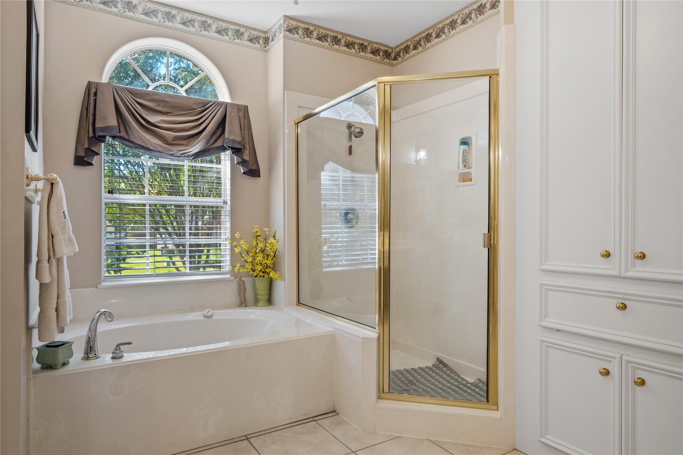 319 Palais Verde Road Montgomery, TX 77356 - Photo 19 of 39 This bathroom features a spacious tub beneath a large window with a decorative arch, allowing natural light to fill the space. Adjacent is a glass-enclosed shower, elegant cabinetry and a neutral tile floor, creating a serene and functional space.