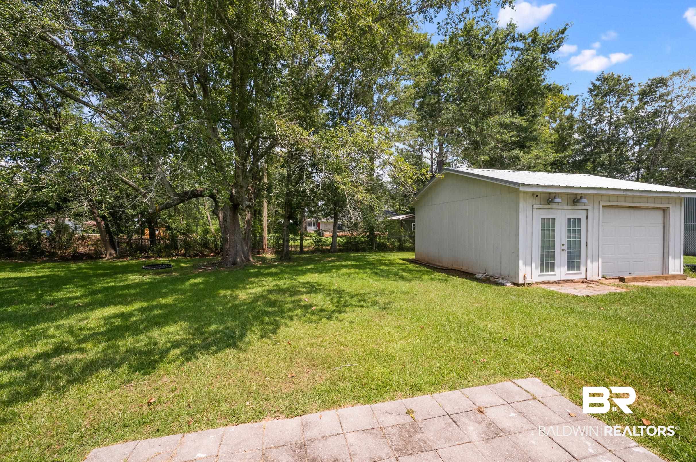24663 Chestnut Street Elberta, AL 36530 - Photo 20 of 22 Expansive backyard featuring a large detached outbuilding with a single garage door and a double glass-paned entry door, a paved patio area, and mature trees providing natural shade