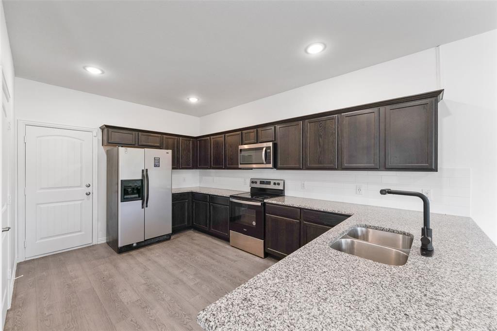 4651 Raleigh Dalton Road Dallas, TX 75227 - Photo 7 of 19 Modern kitchen featuring dark wood cabinetry with glass-front upper doors, stainless steel appliances including a refrigerator and range, quartz countertops with waterfall edge, a deep stainless steel sink with matte black pull-down faucet, recessed ceiling lighting, and wood-finish flooring throughout the open layout