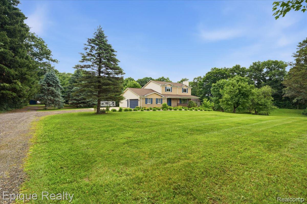 1221 Rhodes Road Lake Orion, MI 48360 - Photo 4 of 72 Expansive green lawn with mature trees and a gravel driveway providing access to a two-story residence featuring a covered front porch and an attached garage