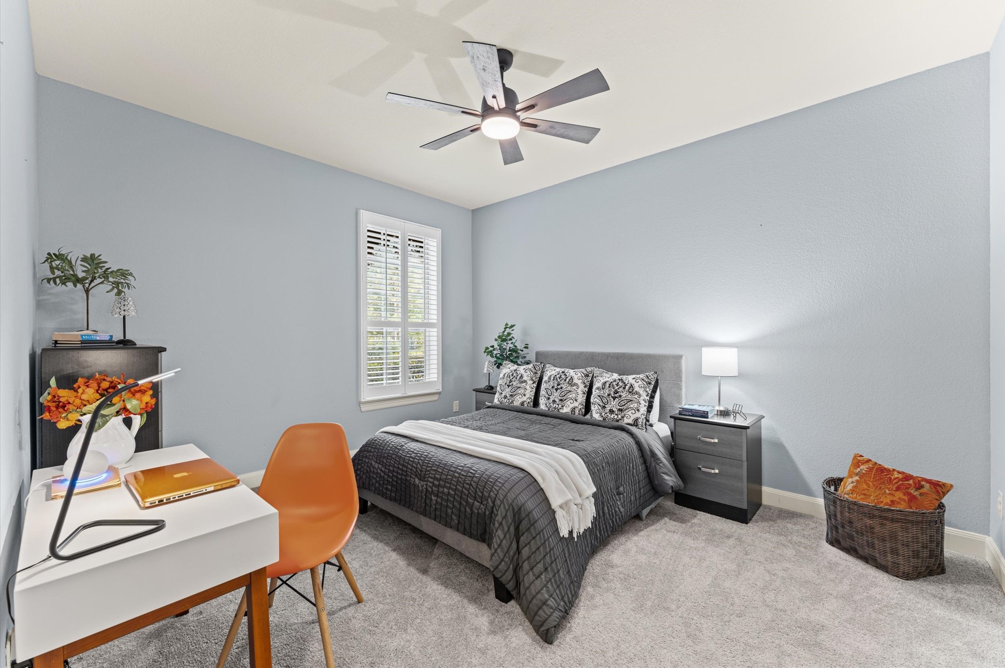 70 Valera Ridge Drive Spring, TX 77389 - Photo 28 of 37 Cozy bedroom with soft blue walls and carpeted floors. Features a modern ceiling fan, a comfortable bed with decorative pillows, and a bedside table with a lamp. A small desk with an orange chair adds functionality, while a window with shutters offers natural light.