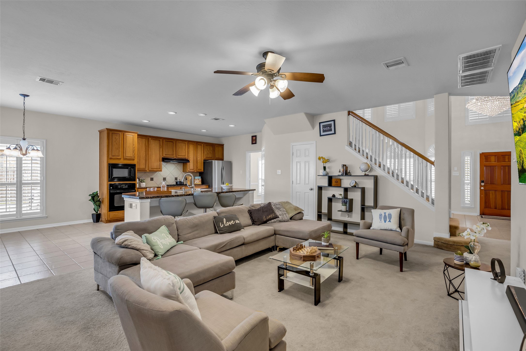 6807 Chapelfield Lane Houston, TX 77049 - Photo 23 of 42 This open-concept living area features a spacious layout with a cozy sectional, modern ceiling fan, and abundant natural light. The kitchen boasts warm wooden cabinetry, a central island with seating, and stainless steel appliances. A staircase leads to the upper level, adding a touch of elegance.