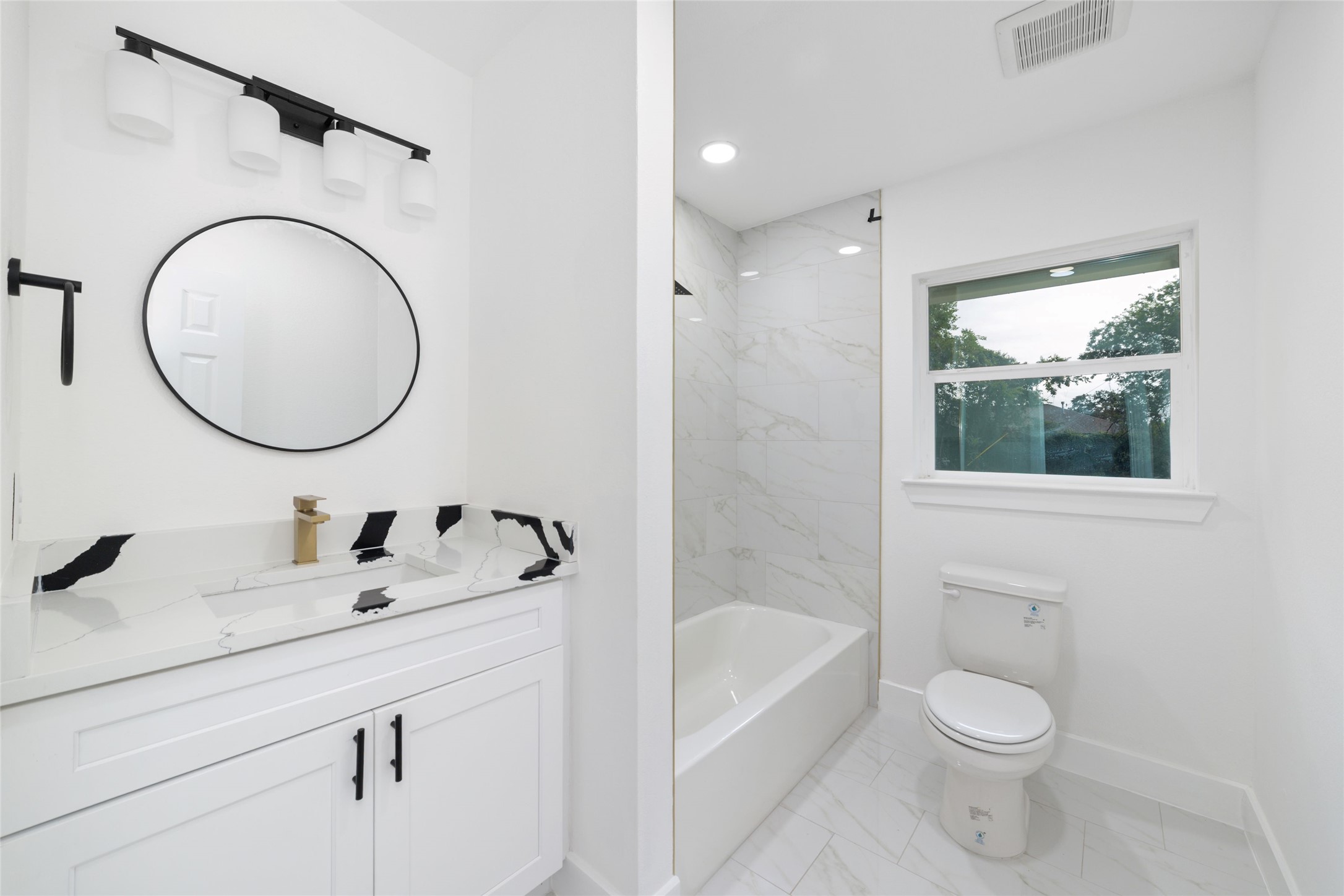 5709 Langley Road Houston, TX 77016 - Photo 19 of 27 This bathroom features a modern design with white cabinetry, a round mirror, and sleek black fixtures. It includes a bathtub with a marble-like tile surround and a window for natural light.