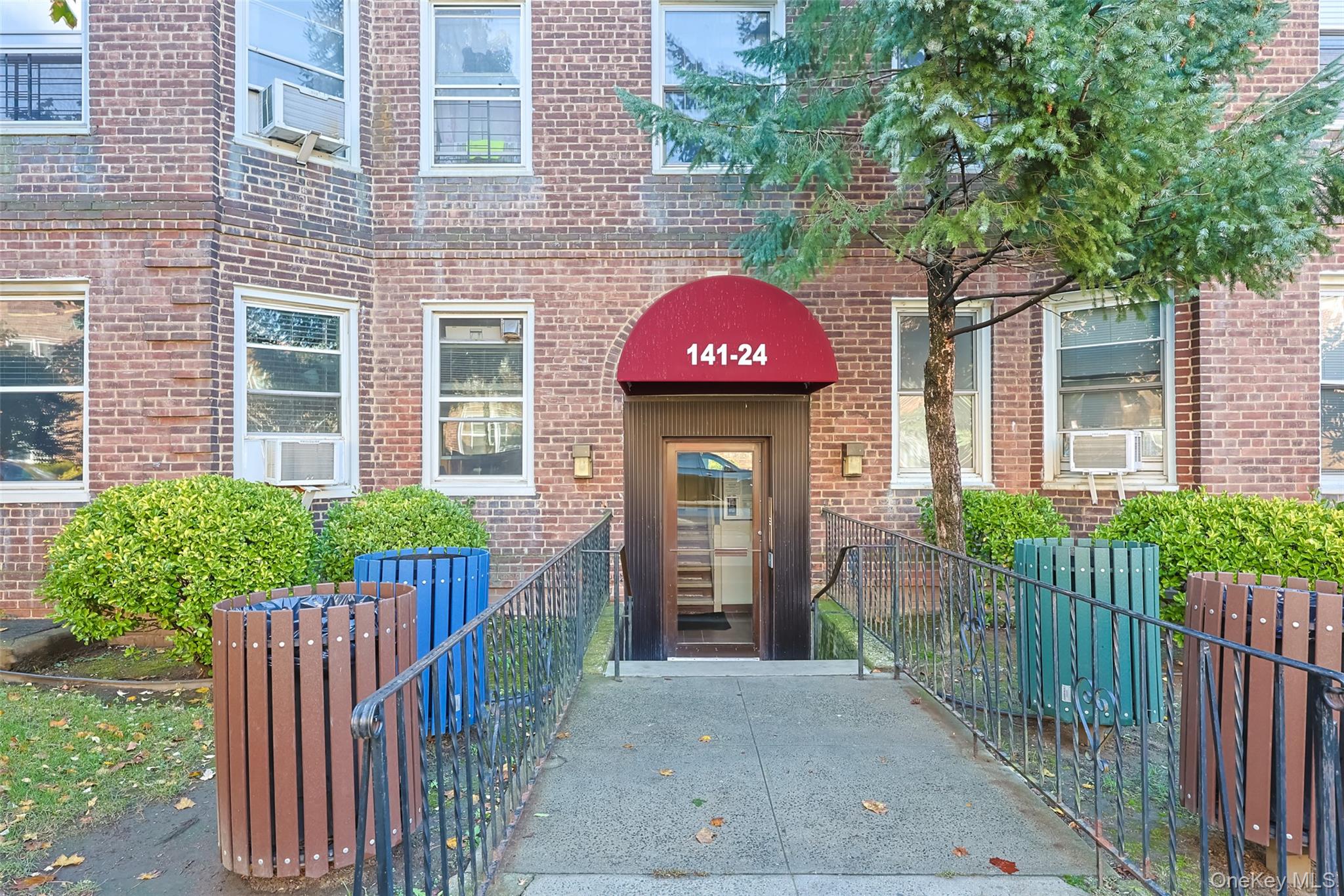 Charming brick building with a welcoming entryway featuring a red canopy and secure glass door access, surrounded by neatly maintained landscaping and mature trees.