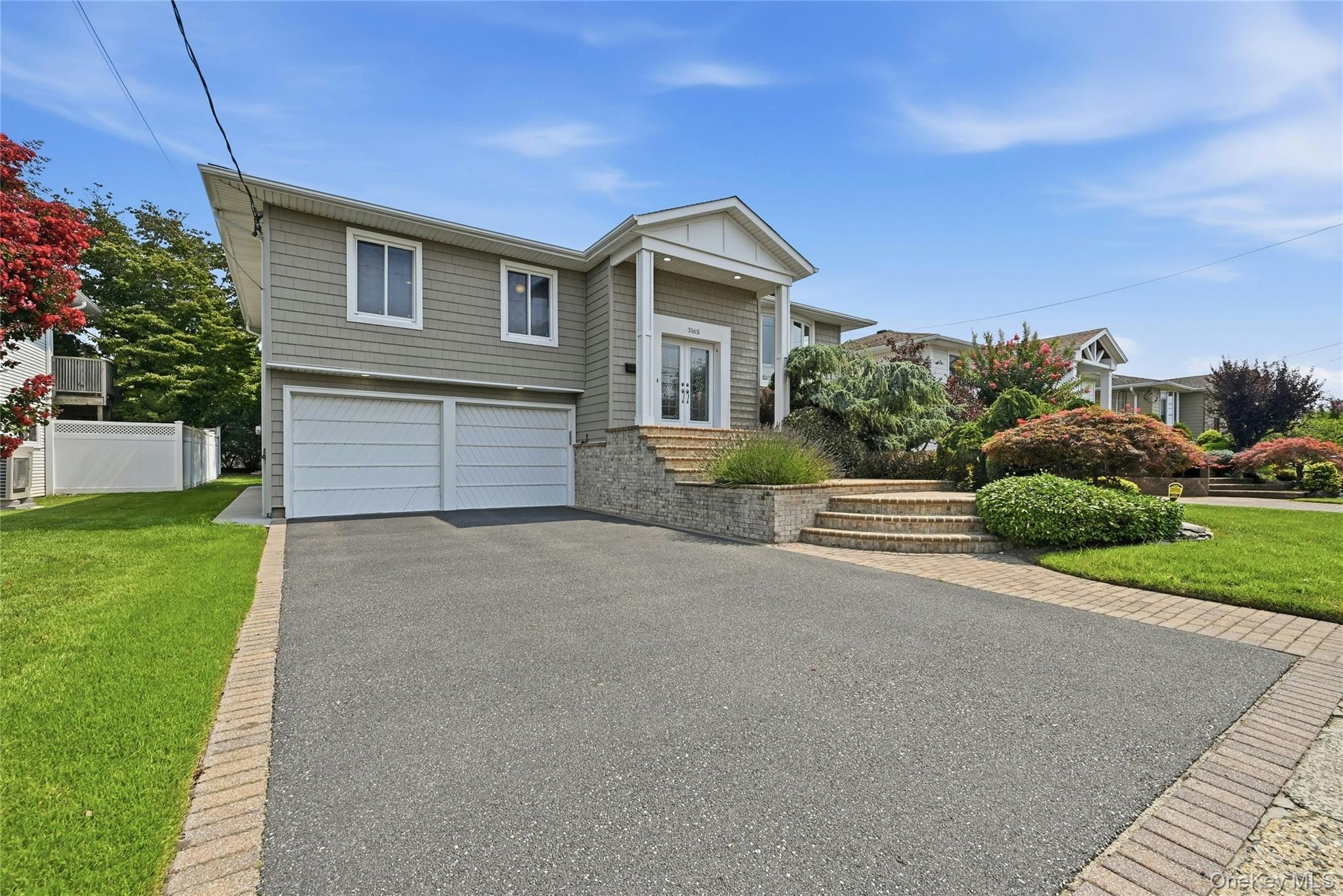 Vinyl siding exterior with multiple windows, two-car garage, tiered stone landscaping, and paved driveway with brick edging