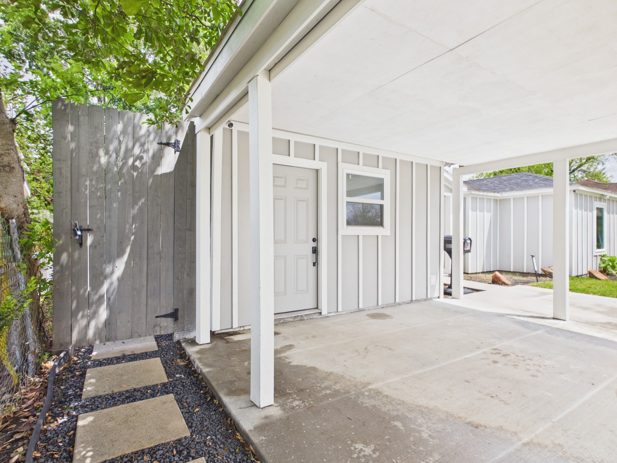 4927 Kelso Street Houston, TX 77021 - Photo 8 of 45 This photo shows a covered carport with a white exterior and vertical siding. It features a side entrance door and a small window. The area includes a paved walkway with stepping stones, and a wooden fence offers privacy.