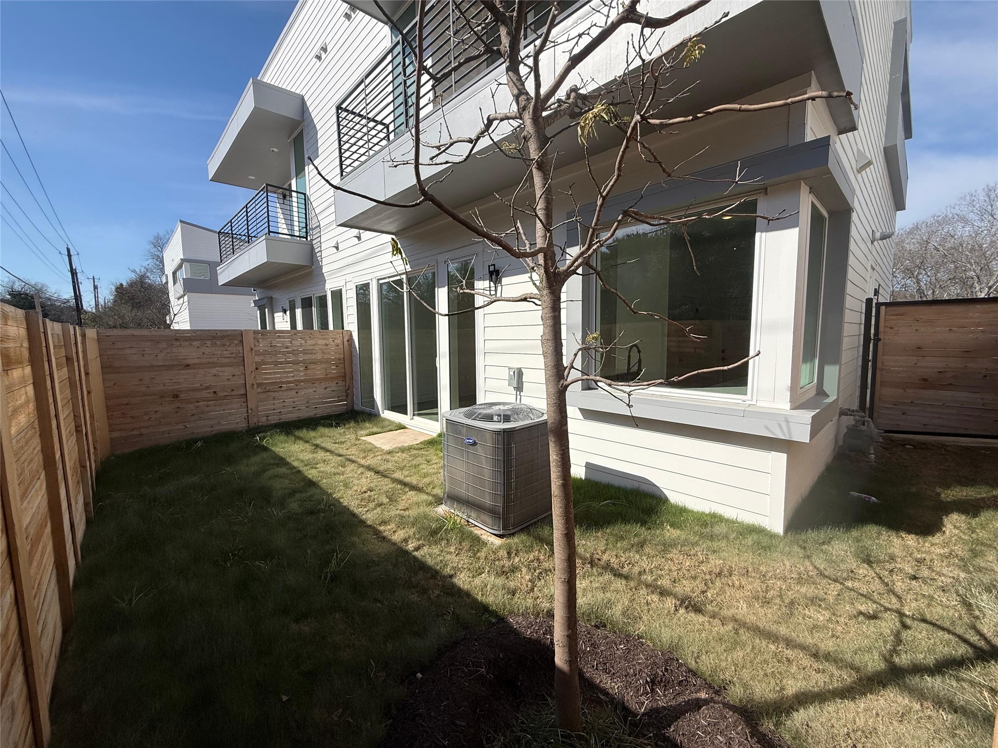 2000 Riverside Farms Road, Unit 8102 Austin, TX 78741 - Photo 9 of 10 This photo shows a modern townhouse exterior featuring a small fenced backyard with a young tree and an HVAC unit. The building has large windows and balconies, providing a bright and airy feel. The yard offers a cozy outdoor space for relaxation or gardening.