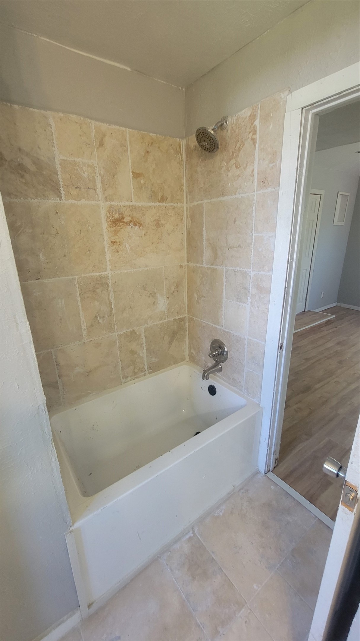 12018 Sandpiper Road Angleton, TX 77515 - Photo 15 of 48 This photo shows a bathroom with a tiled shower and bathtub combination. The walls are lined with beige tiles, and the floor has matching tiles. A single showerhead is installed above the tub. The entrance to the bathroom opens to a room with wood-style flooring.
