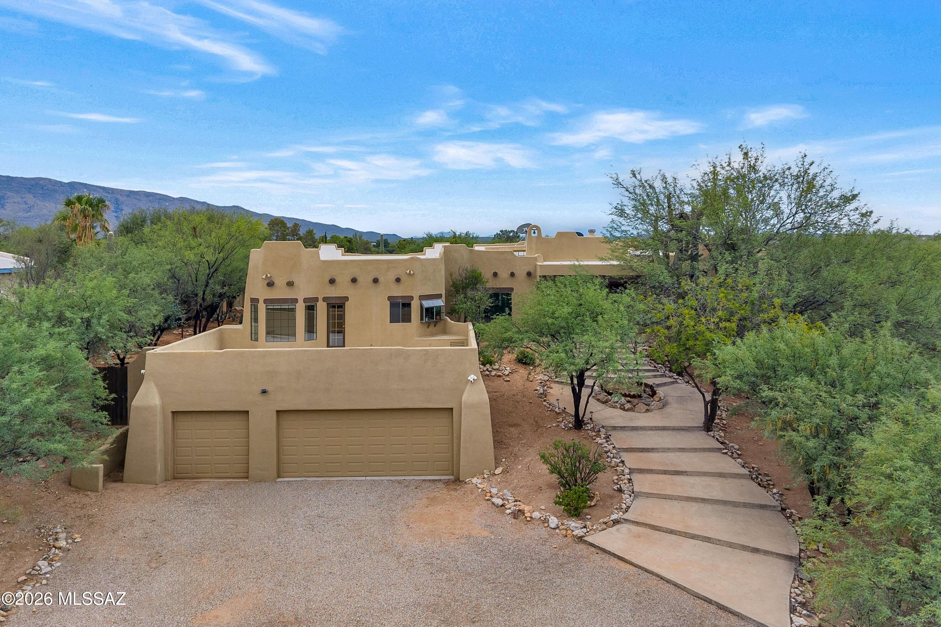 12160 East Tanque Verde Road Tucson, AZ 85749 - Photo 1 of 53 Southwestern style home with an attached three-car garage, gravel driveway, and concrete walkway framed by mesquite trees and Santa Catalina Mountain views.