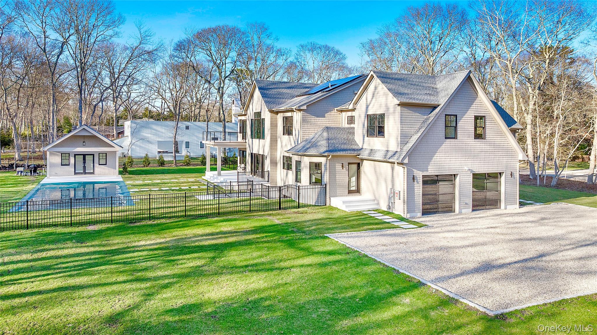 Contemporary shaker-shingle residence featuring a two-car garage with frosted glass doors, an expansive gravel driveway, and a fenced-in swimming pool with a pool house, with abutting creek front nature preserve.