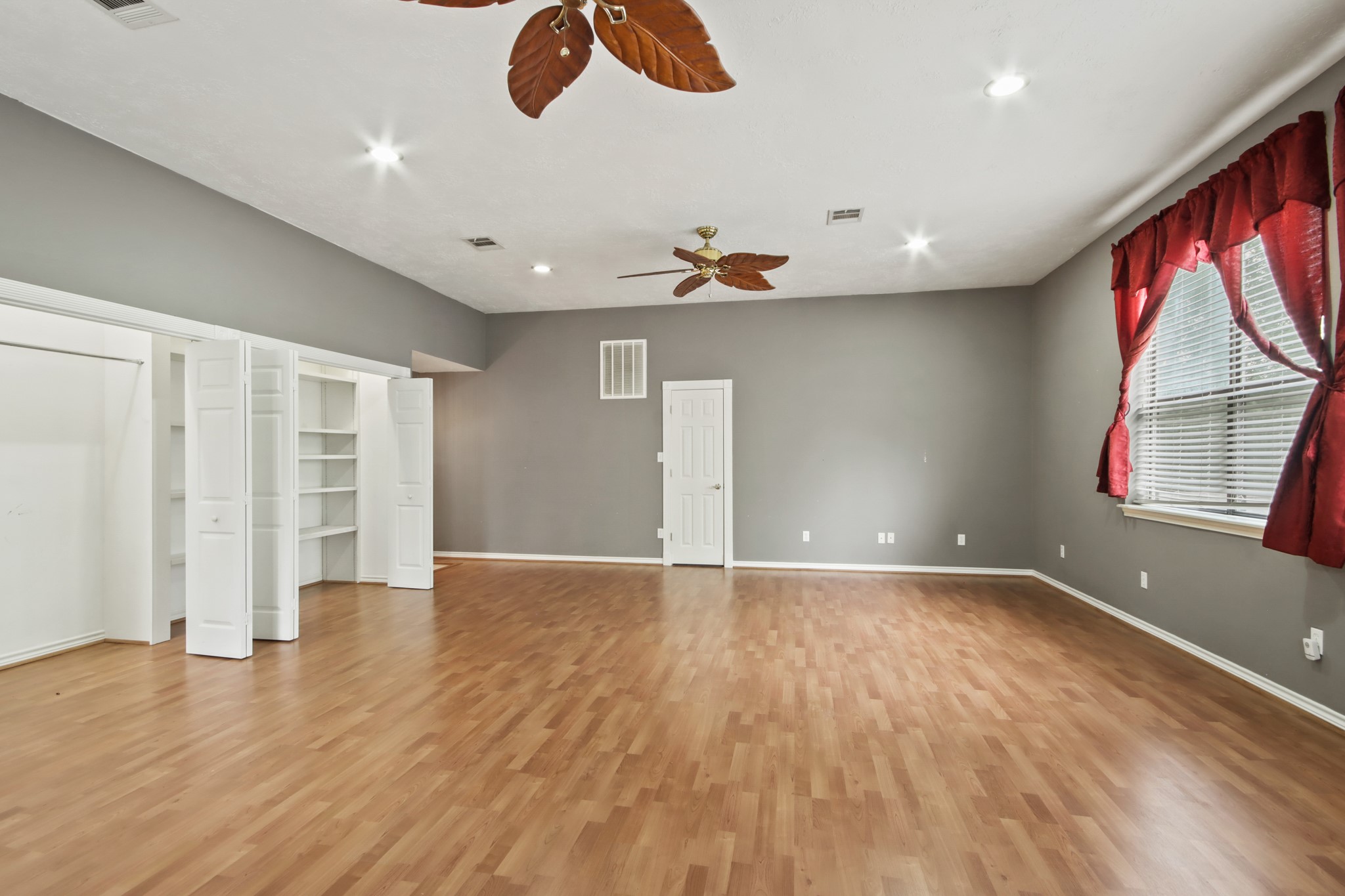 35 Susan Drive Cleveland, TX 77327 - Photo 18 of 33 This spacious room features light wood flooring, neutral gray walls, and ample natural light from large windows with red curtains. It includes built-in shelving, a closet with double doors, and ceiling fans for comfort.