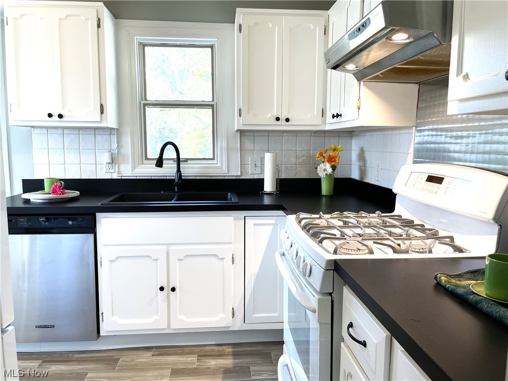 7849 State Road Cleveland, OH 44134 - Photo 7 of 34 Kitchen has a white gas range, stainless steel dishwasher, wall chimney exhaust hood, dark countertops, white cabinets, new wood-type laminate flooring, and backsplash.