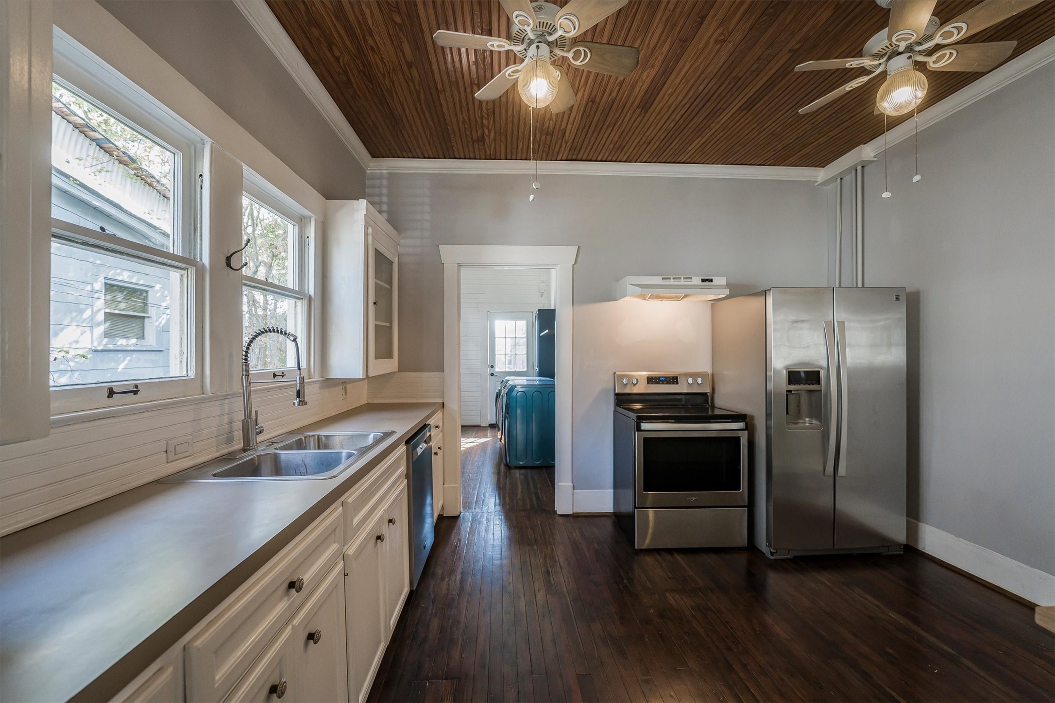210 Tabor Street Houston, TX 77009 - Photo 6 of 18 This kitchen features modern stainless steel appliances, including a refrigerator and oven, with sleek white cabinetry and a double sink. The room boasts a wood-paneled ceiling with ceiling fans and warm wood flooring, creating a cozy, inviting atmosphere.