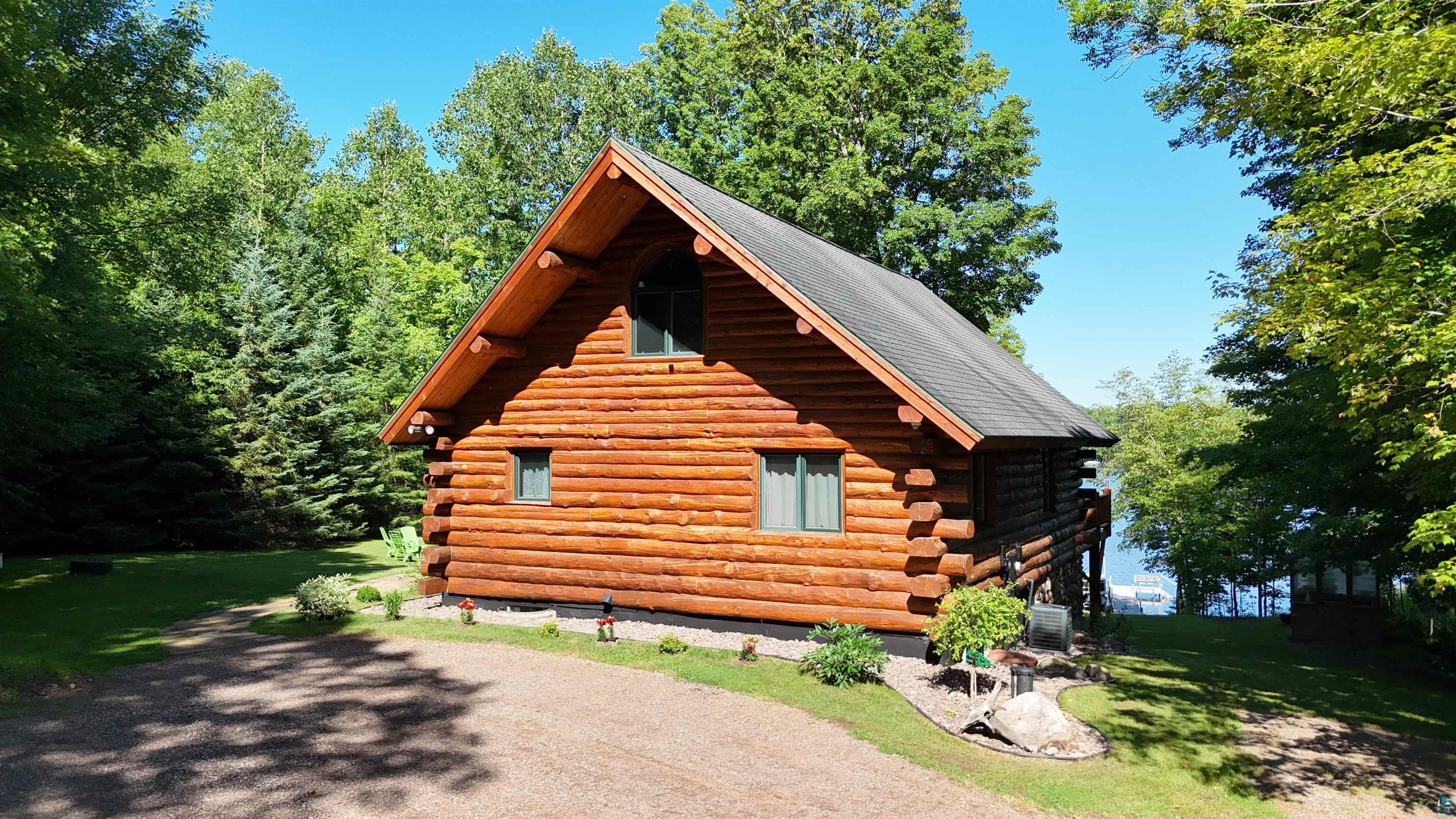 4613 North Lake Winter Road Winter, WI 54896 - Photo 68 of 73 Log cabin exterior with a pitched roof, surrounded by mature trees, a gravel driveway, and a visible body of water