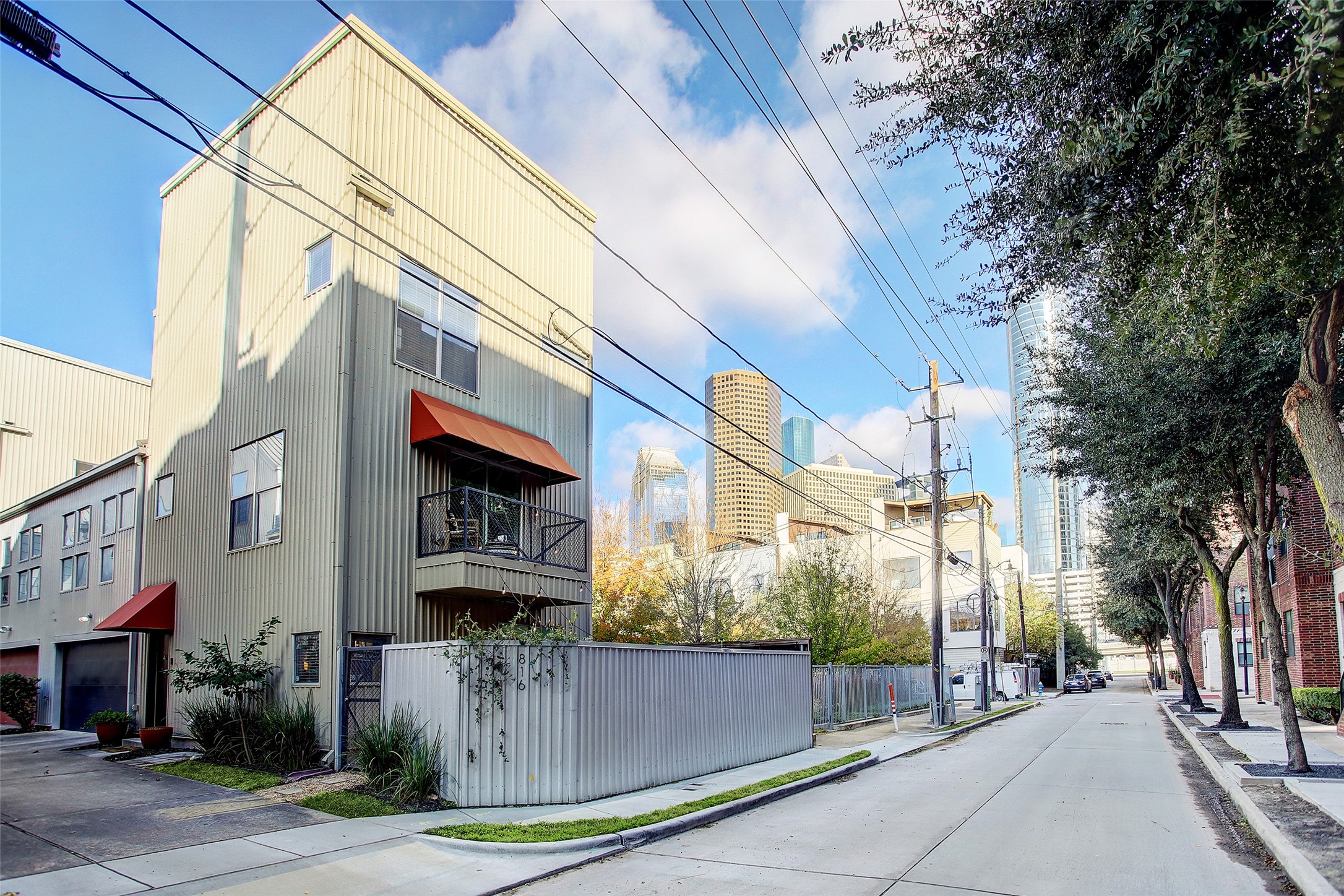 Modern, multi-story townhome with metal siding and red awnings, featuring a small balcony. Located in an urban setting with a view of downtown skyscrapers. The street is lined with trees and offers a clean, residential feel.